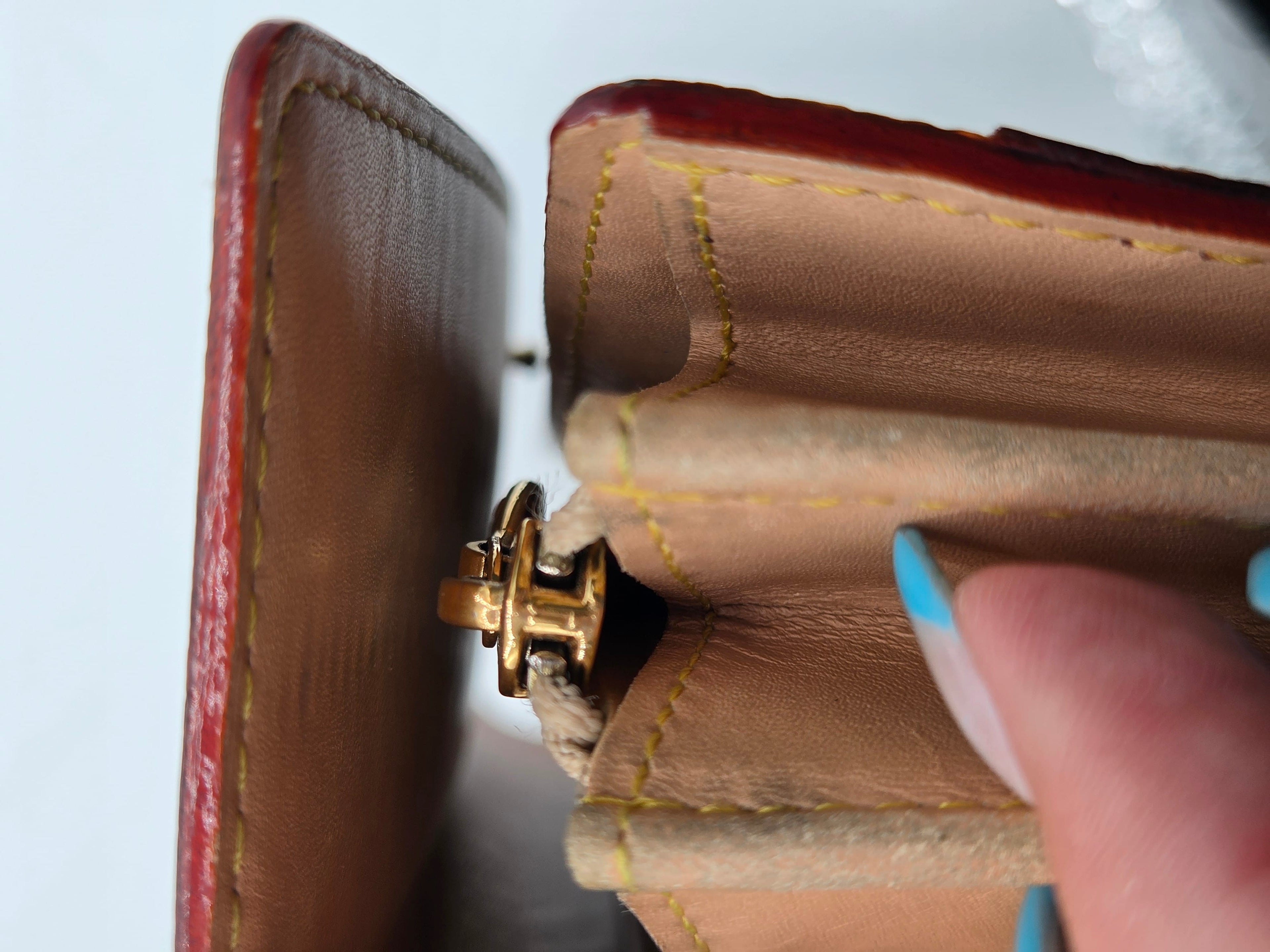 Close-up of an open brown leather wallet or card case showing tan interior lining, yellow stitching, a gold-tone clasp, and a finger with blue nail polish touching the edge.