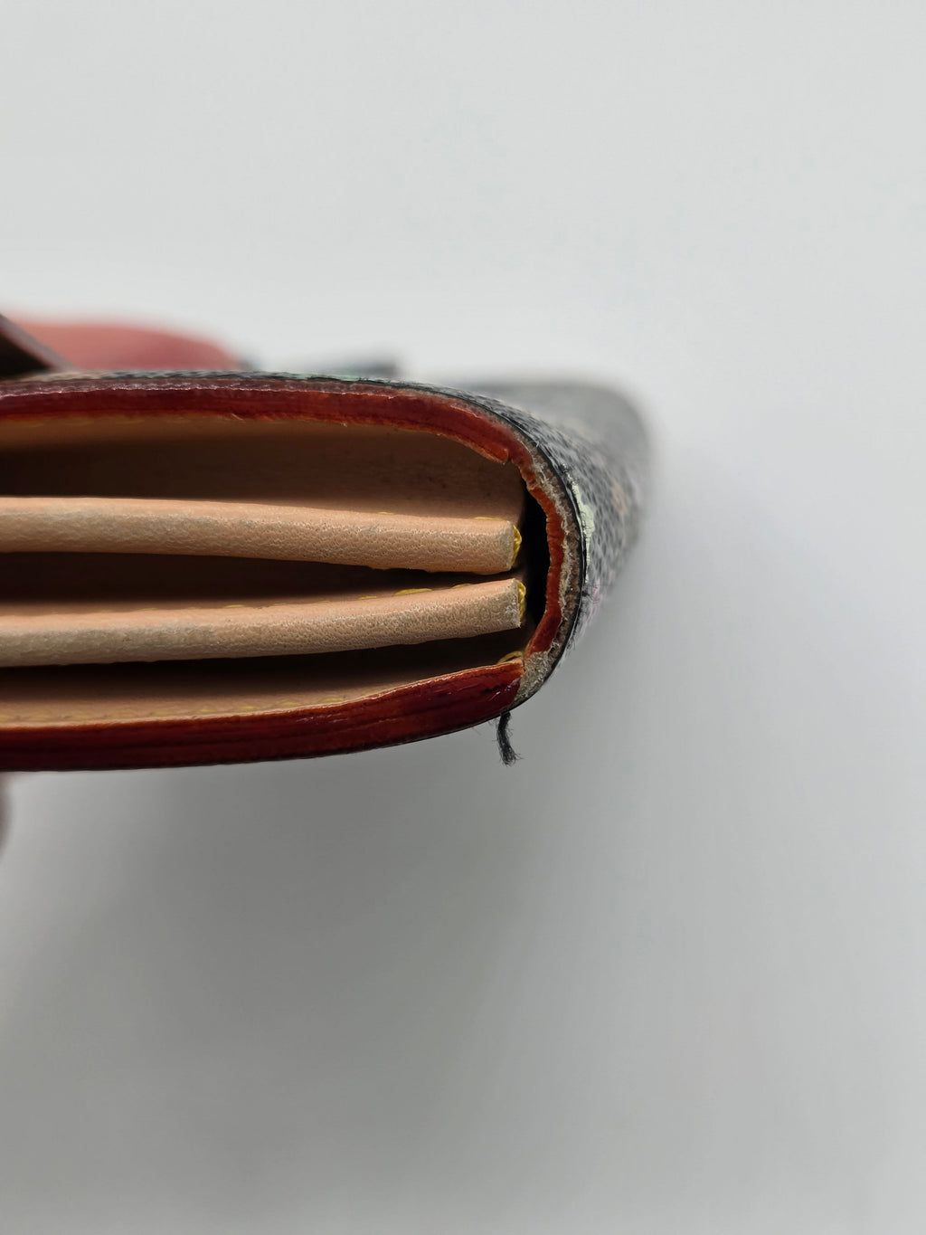 Close-up of the edge of a leather notebook or journal showing the dark textured cover, reddish-brown interior lining, and light tan pages against a plain light gray background.
