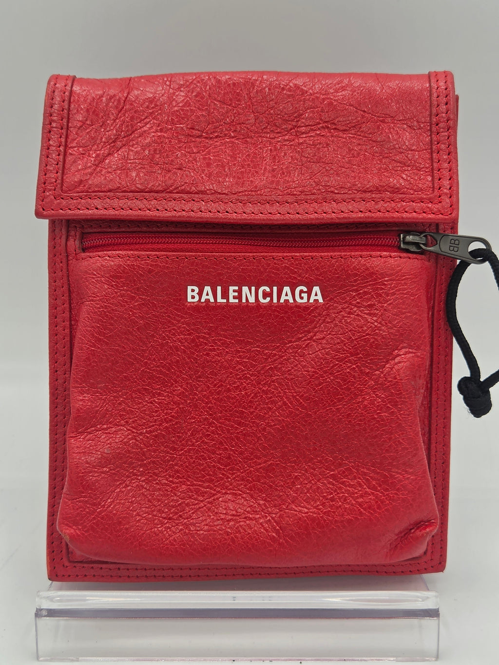 A red Balenciaga leather zippered pouch with a vertical white BALENCIAGA logo, shown on a light gray background next to a clear plastic container.