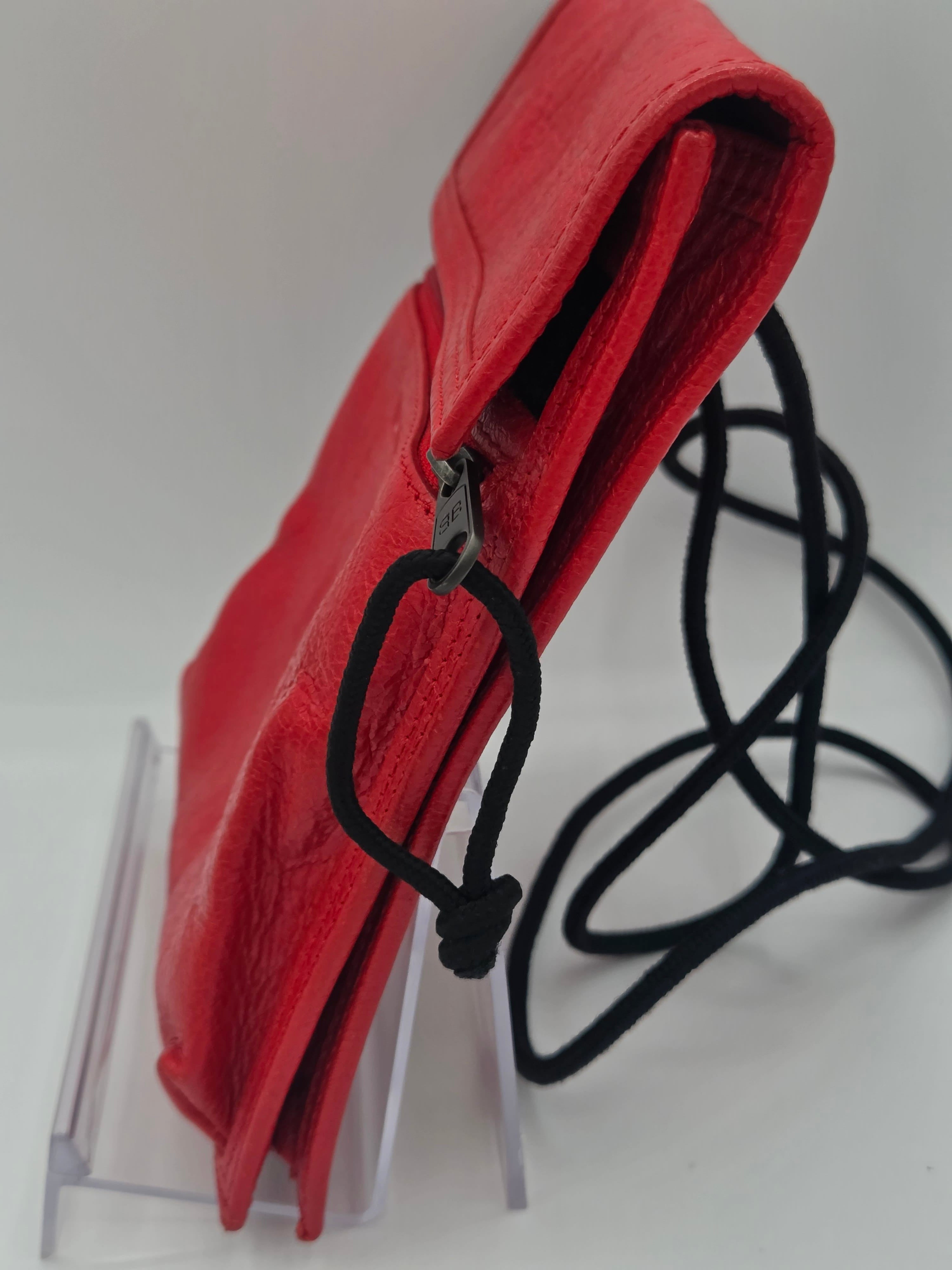 A red leather pouch with a black drawstring cord and a silver metal clasp, displayed on a clear acrylic stand against a white background.