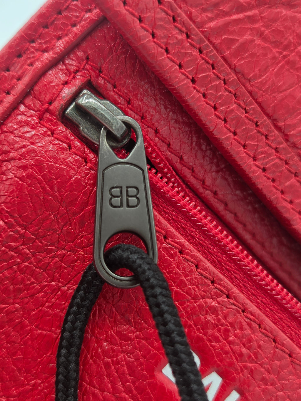 Close-up of a red textured leather zipper with a dark gray metal pull tab engraved with 'BB' and a black braided cord passing through the ring.