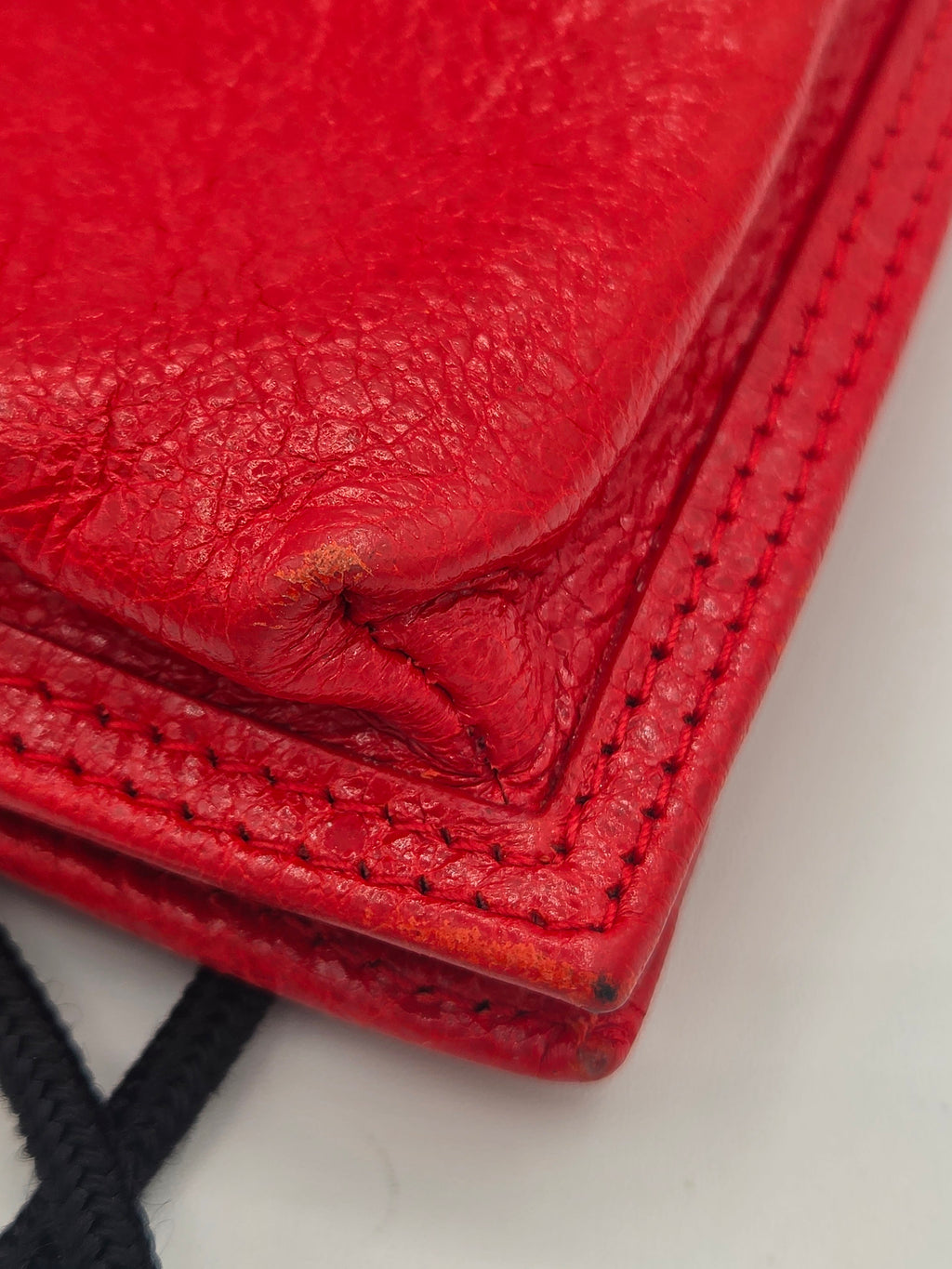 Close-up of a bright red textured leather wallet corner with visible stitching and a black strap in the background.