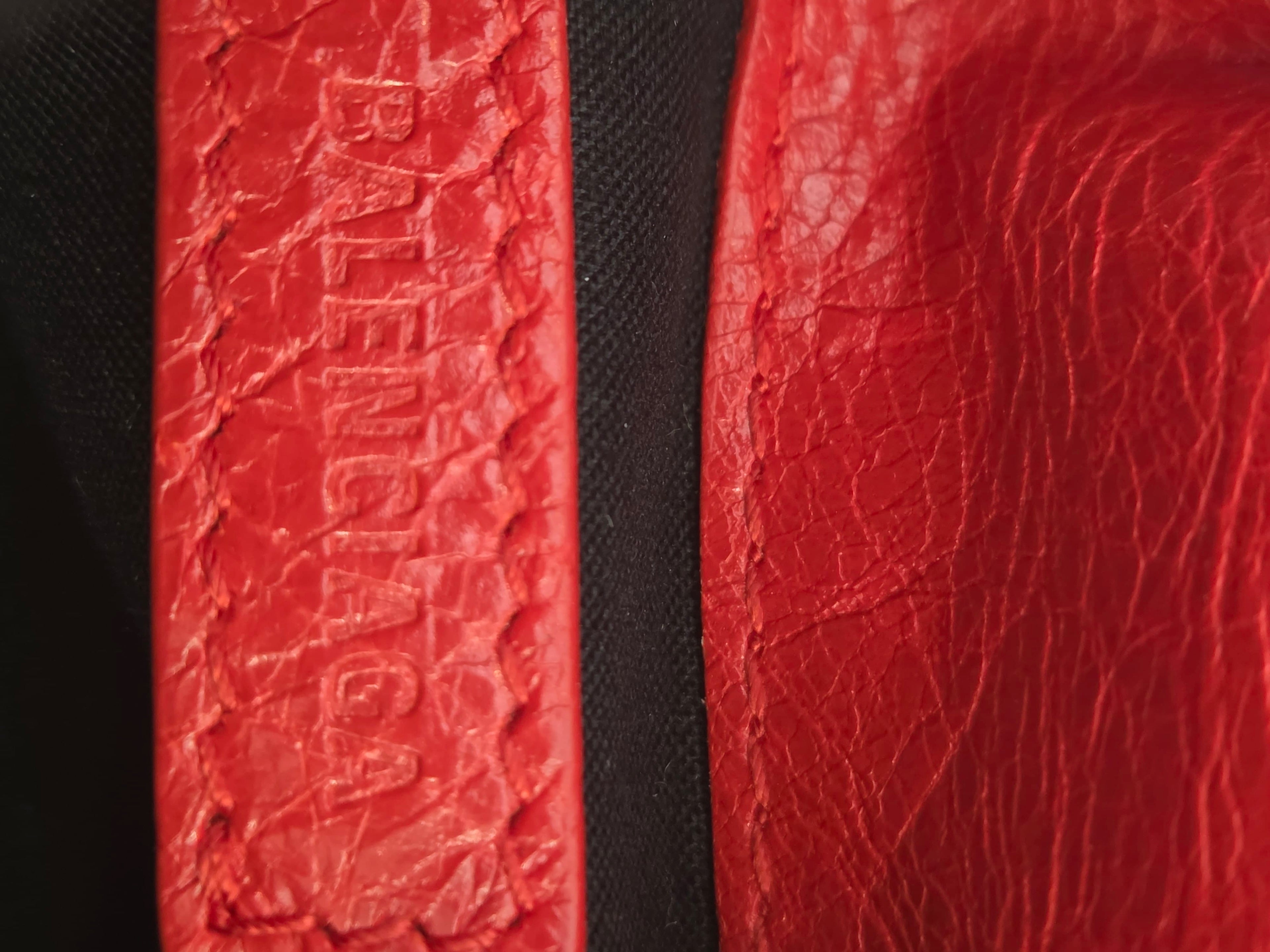 Close-up of a red textured leather strap with embossed Balenciaga branding, shown next to a black fabric background.