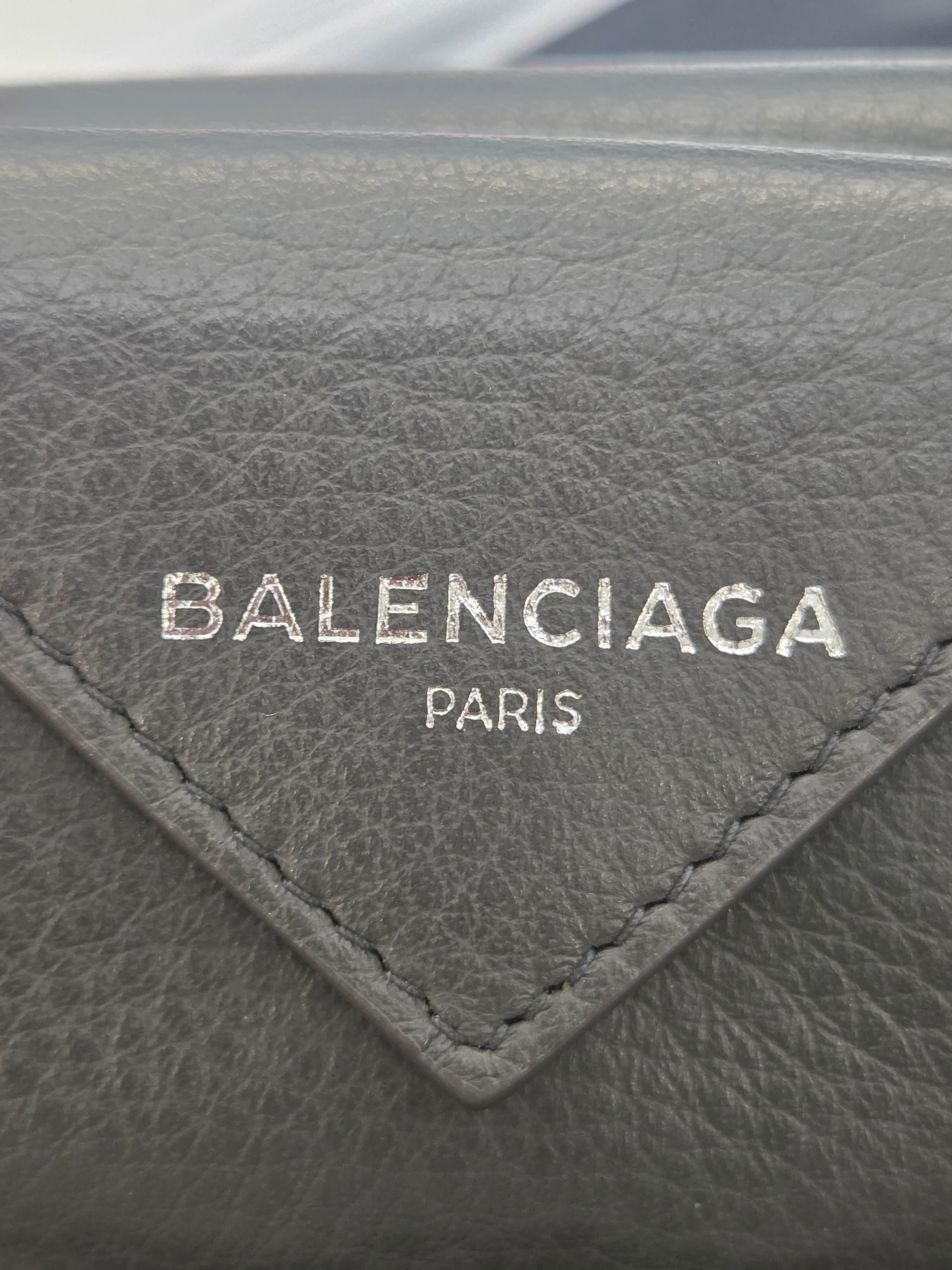 Close-up of a dark gray textured leather item featuring a triangular stitched patch with the Balenciaga Paris logo in silver-tone lettering.