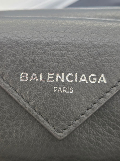 Close-up of a dark gray textured leather item featuring a triangular stitched patch with the Balenciaga Paris logo in silver-tone lettering.
