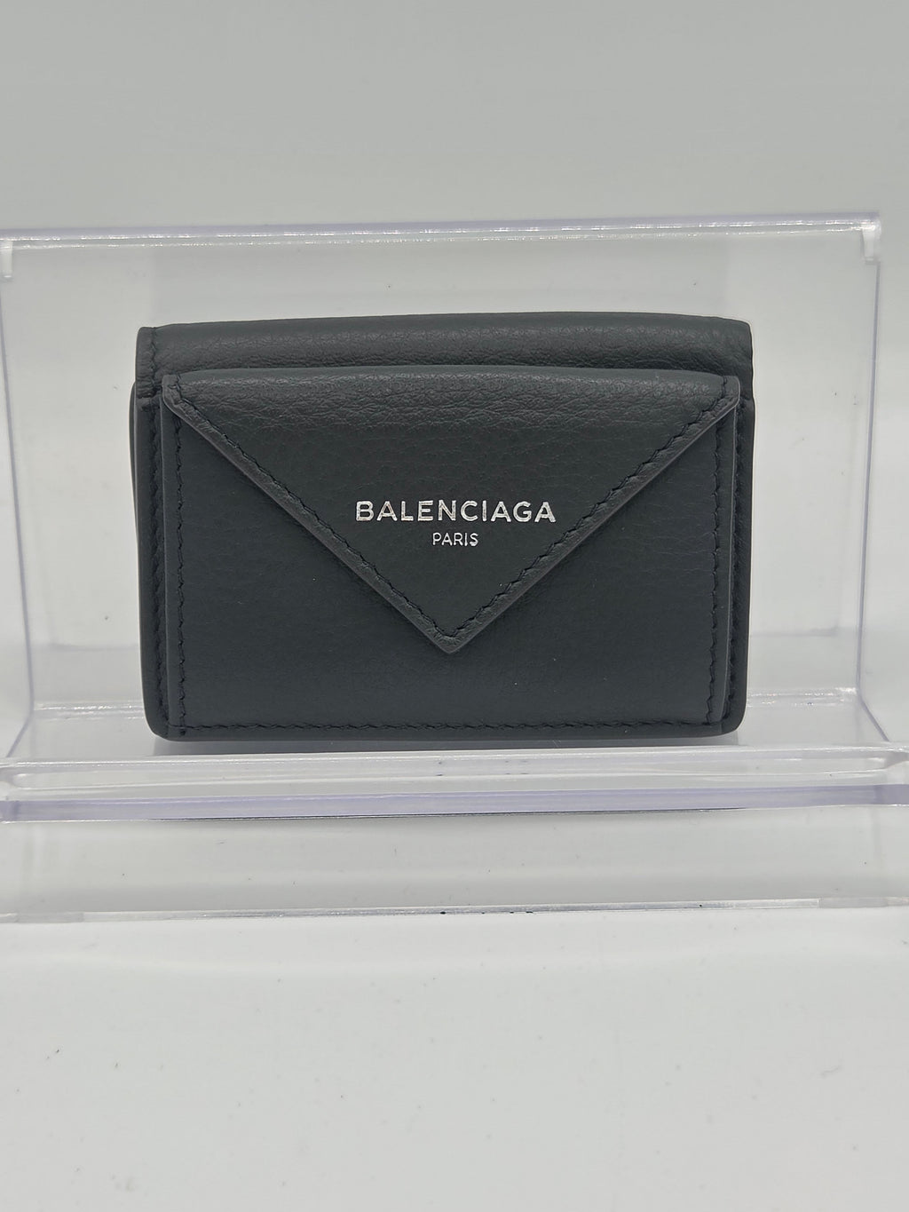 A dark gray Balenciaga leather wallet displayed inside a clear acrylic block on a light gray background.