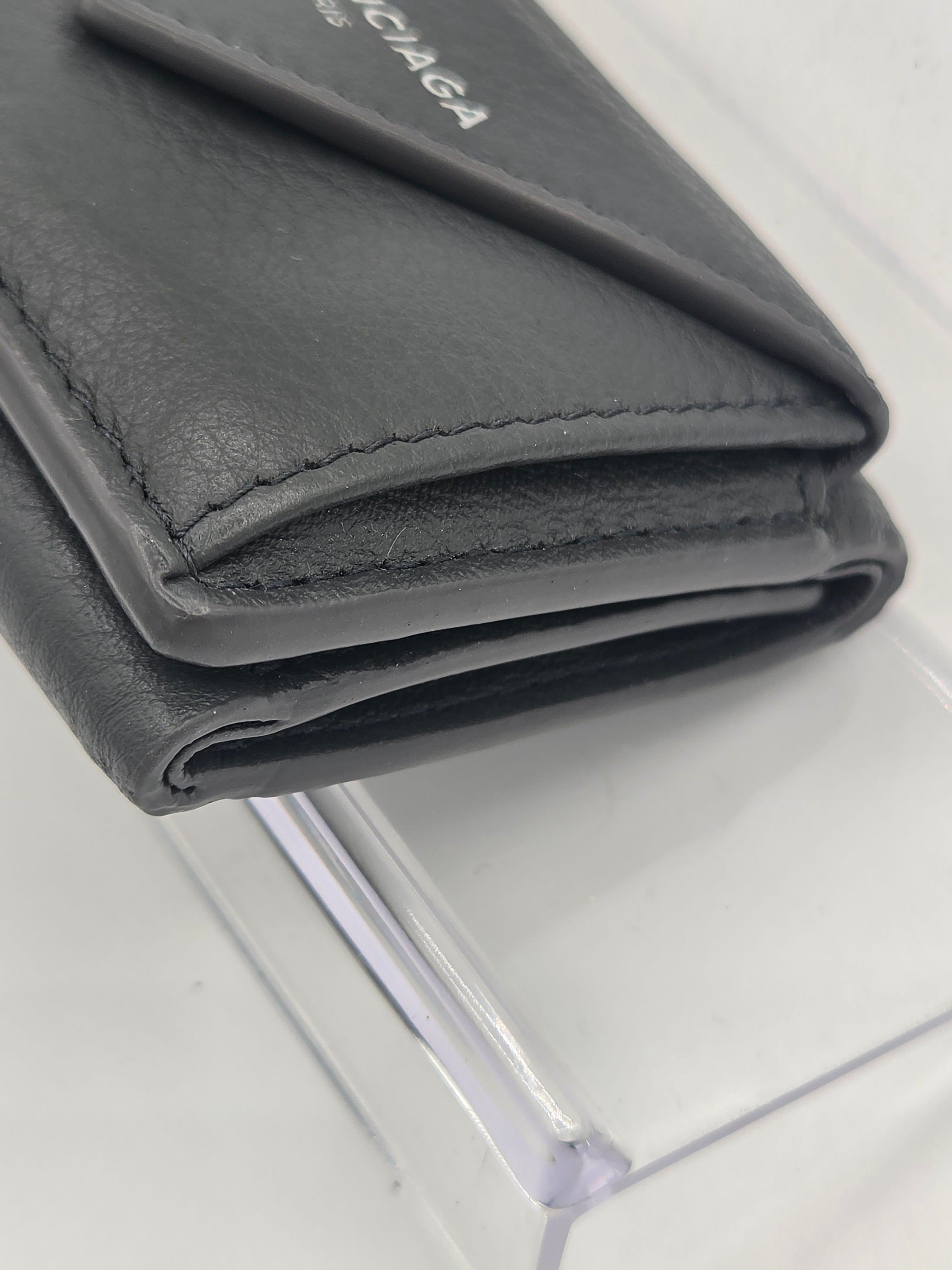 A close-up of a dark gray leather wallet showing the textured grain, stitching, and a portion of the white Balenciaga logo on the side, resting on a clear acrylic block.