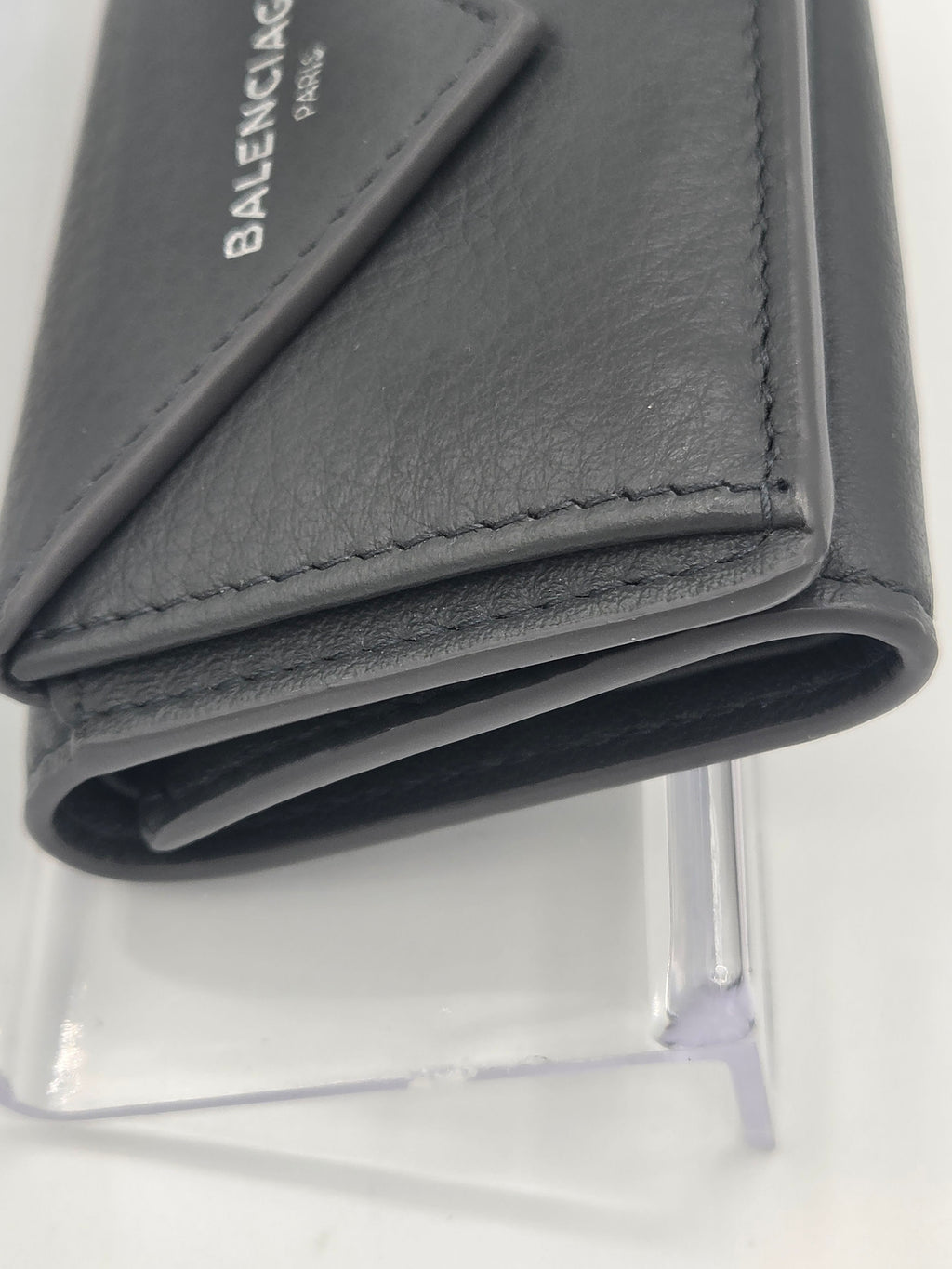 A close-up of a dark gray Balenciaga leather wallet or card holder, showing the textured leather, stitching, and the white Balenciaga logo on the front, displayed on a clear stand against a white background.