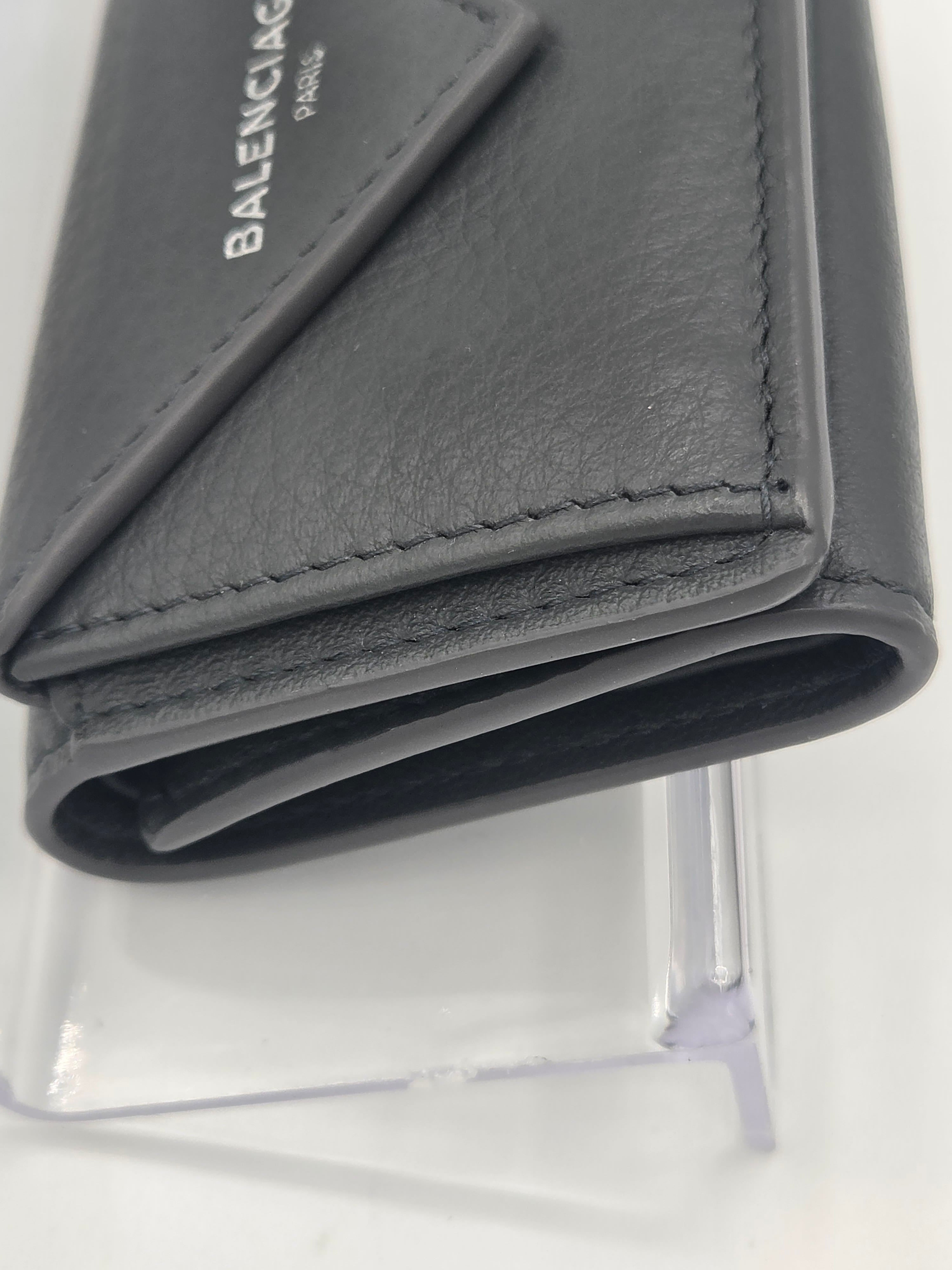 A close-up of a dark gray Balenciaga leather wallet or card holder, showing the textured leather, stitching, and the white Balenciaga logo on the front, displayed on a clear stand against a white background.