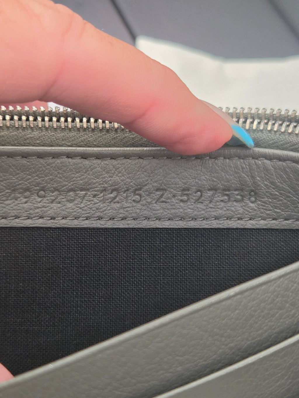 Close-up of a gray textured leather zipper pull with embossed serial numbers, held by a hand with a light blue manicured fingernail next to a black fabric panel.