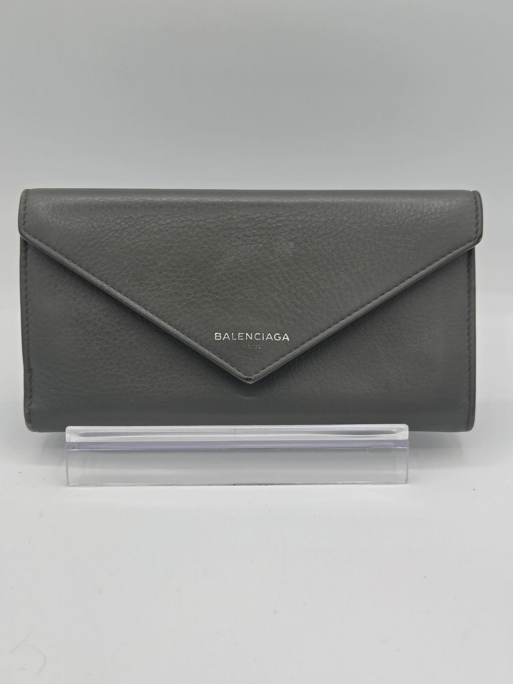 A dark gray textured leather Balenciaga envelope wallet standing on a clear acrylic display block against a neutral gray background.