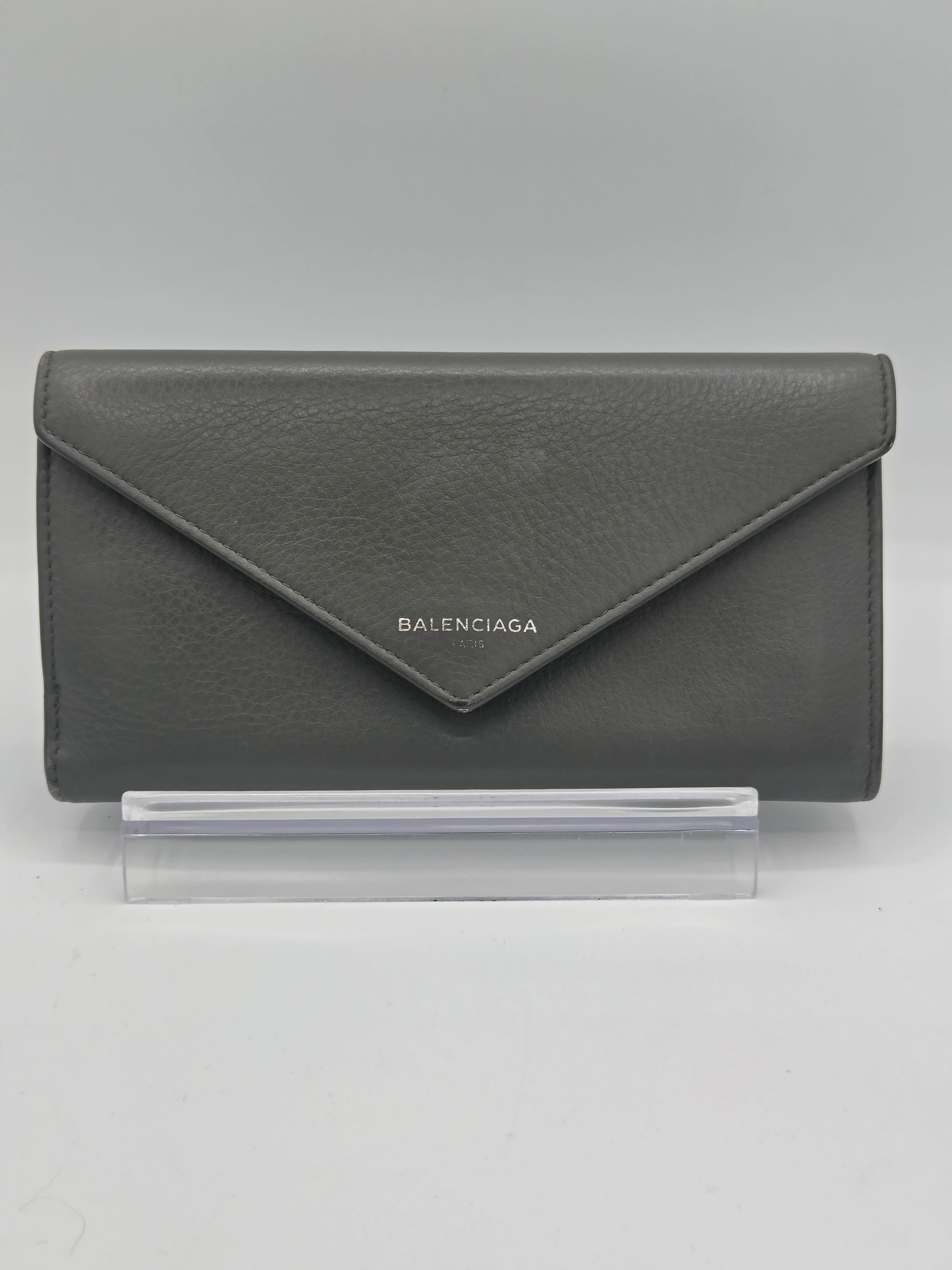 A dark gray textured leather Balenciaga envelope wallet standing on a clear acrylic display block against a neutral gray background.