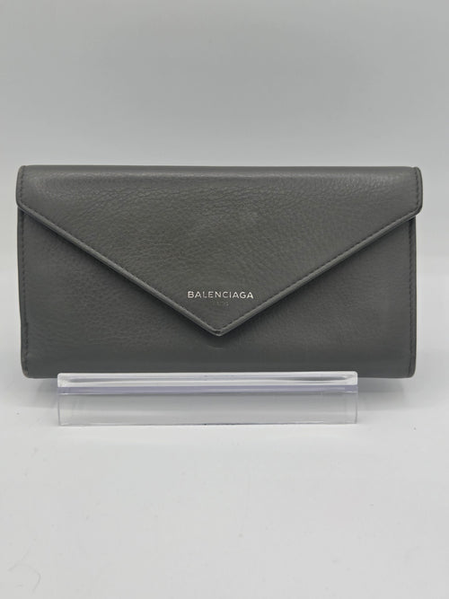 A dark gray textured leather Balenciaga envelope wallet standing on a clear acrylic display block against a neutral gray background.