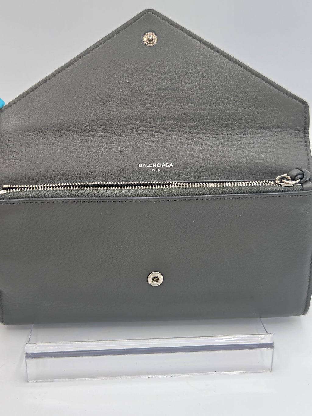 A gray pebbled leather Balenciaga wallet shown open, displaying its interior and silver-tone hardware, positioned next to a clear plastic sleeve on a white background.