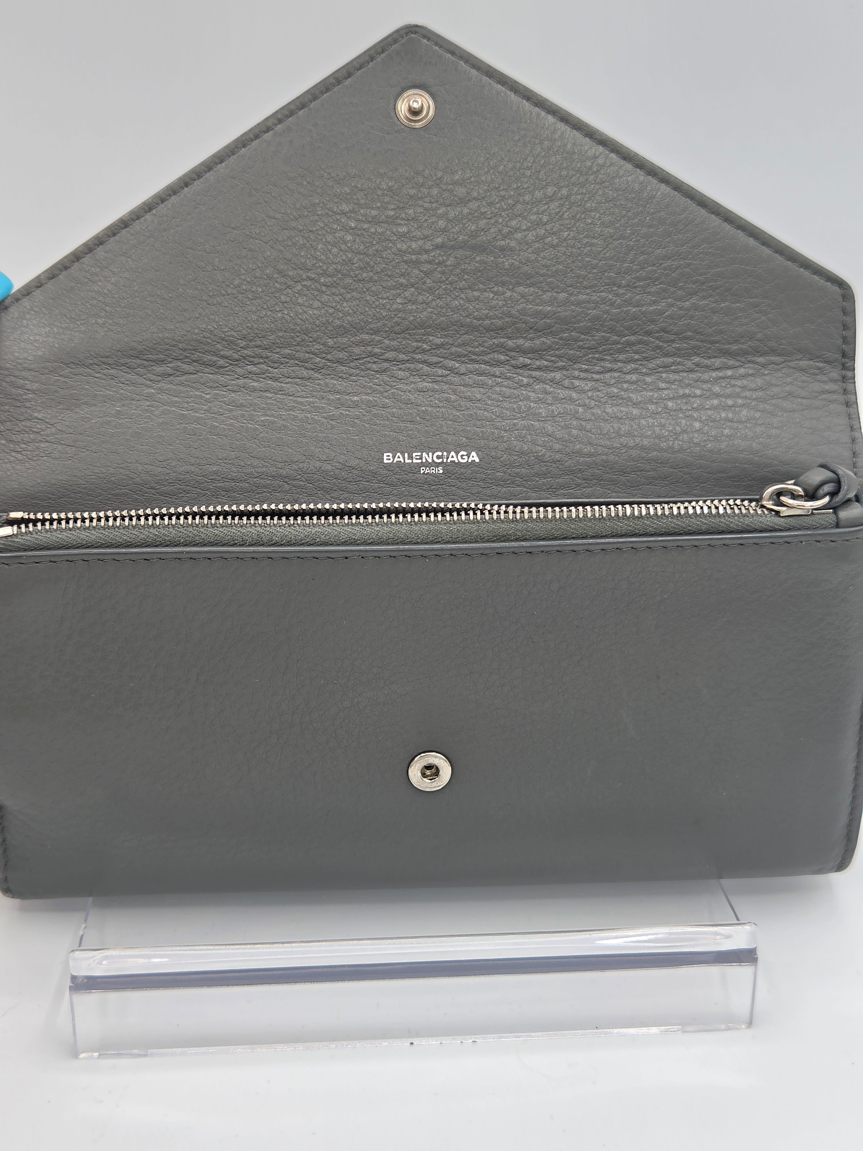 A gray pebbled leather Balenciaga wallet shown open, displaying its interior and silver-tone hardware, positioned next to a clear plastic sleeve on a white background.