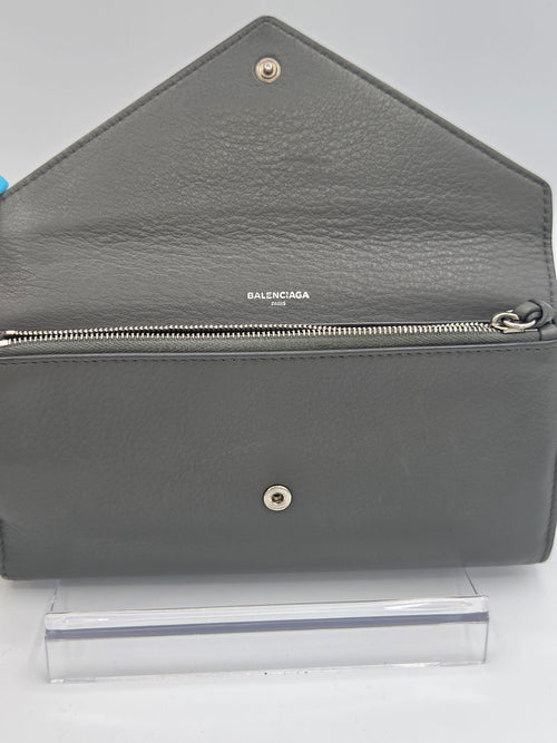 A gray pebbled leather Balenciaga wallet shown open, displaying its interior and silver-tone hardware, positioned next to a clear plastic sleeve on a white background.