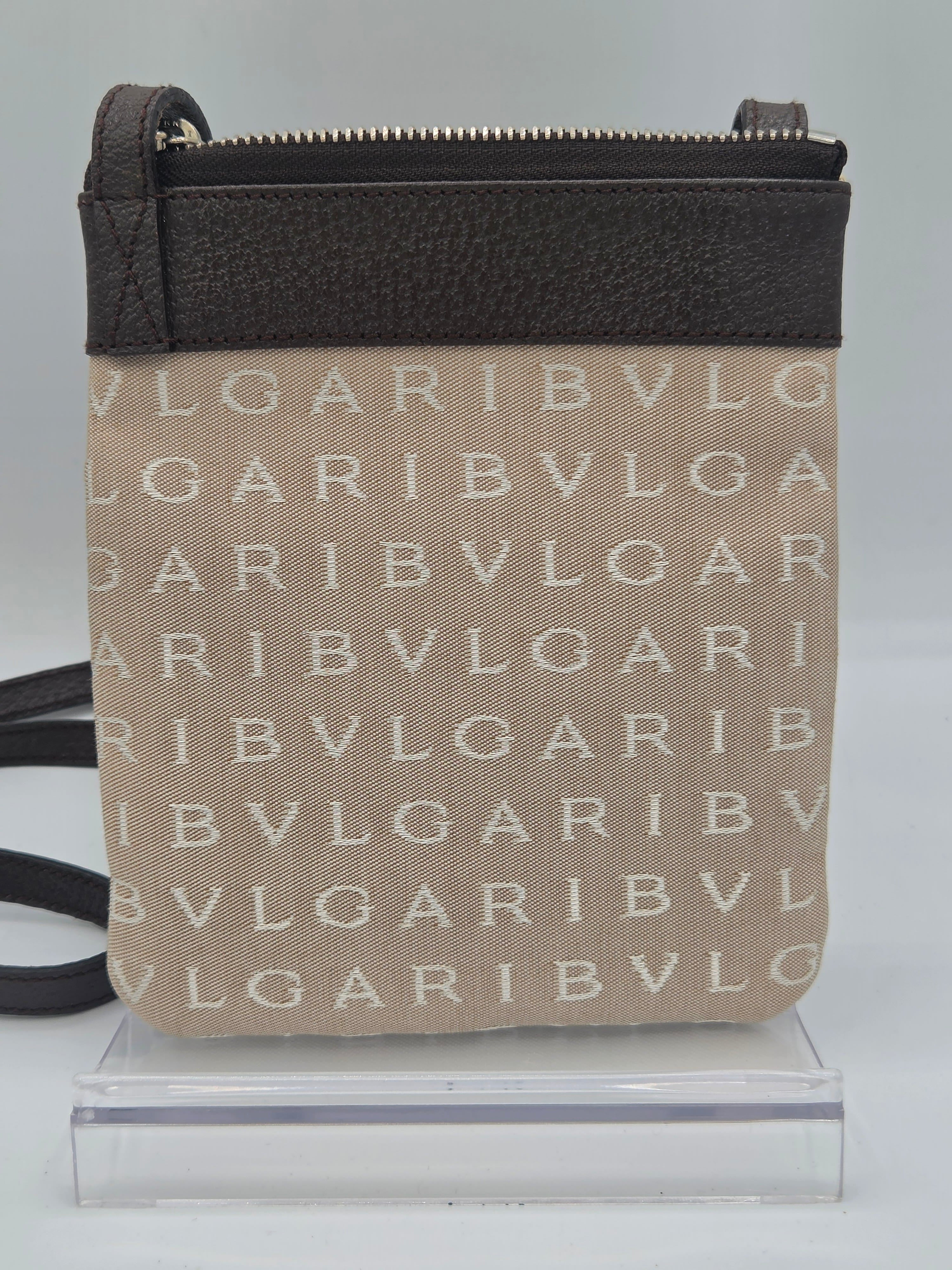 A small crossbody bag with a beige canvas body featuring a repeating white BVLGARI logo pattern, a dark brown textured leather top panel, and a black strap, displayed on a clear acrylic stand against a light gray background.