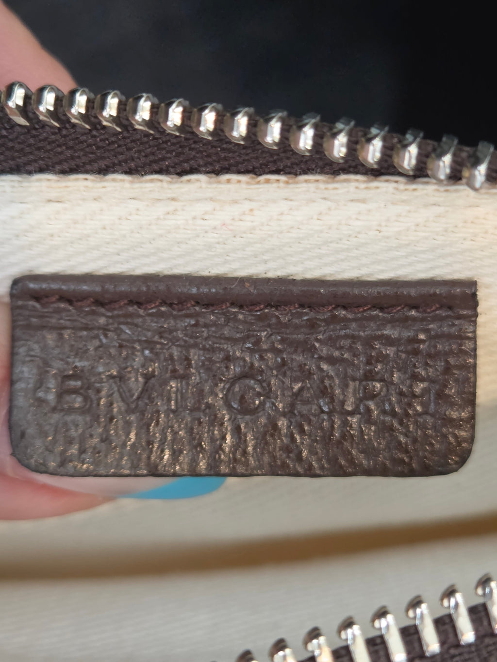 A close-up of a dark brown textured leather patch with embossed lettering, held between fingers next to a cream canvas material and silver zippers.