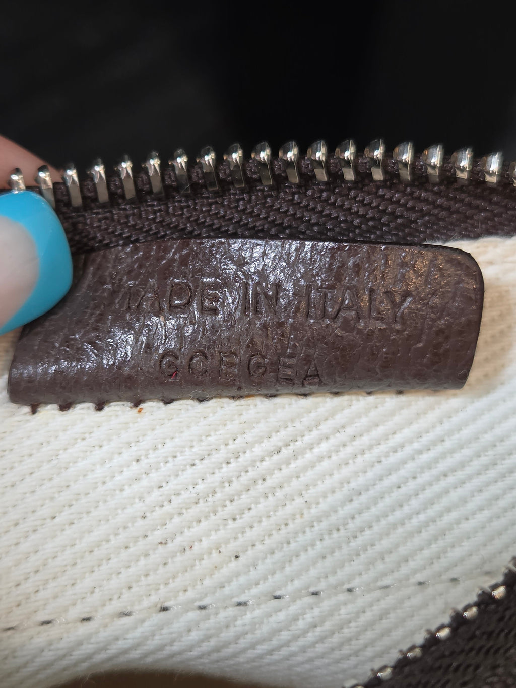Close-up of a dark brown leather zipper pull tab with embossed text reading 'Made in Italy, S.Cecca' next to a white woven textile and silver zipper teeth.