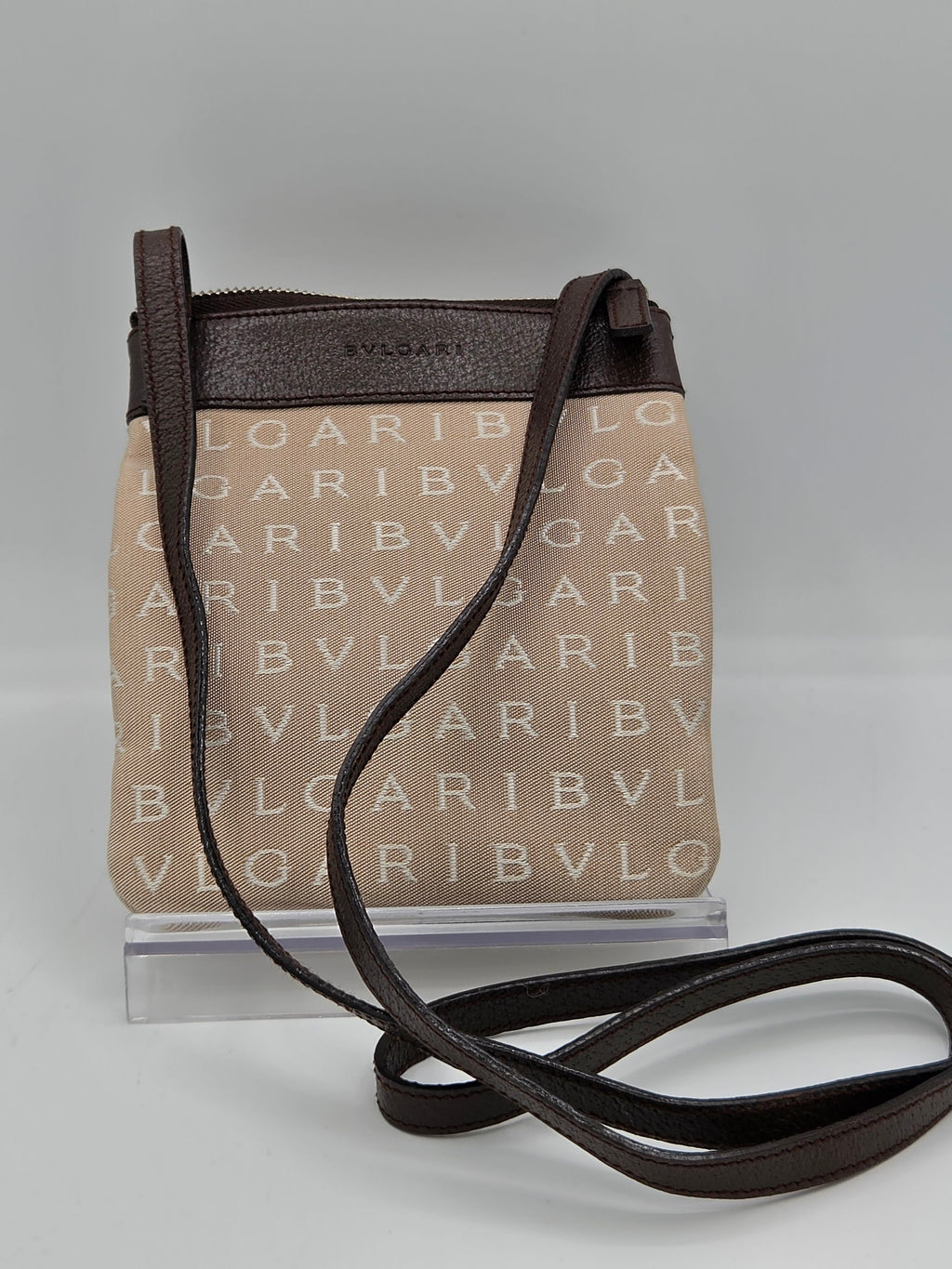 A small crossbody bag with a beige canvas body featuring a repeating 'BVLGARI' monogram pattern, dark brown leather trim and strap, and a clear plastic display insert on a light gray background.