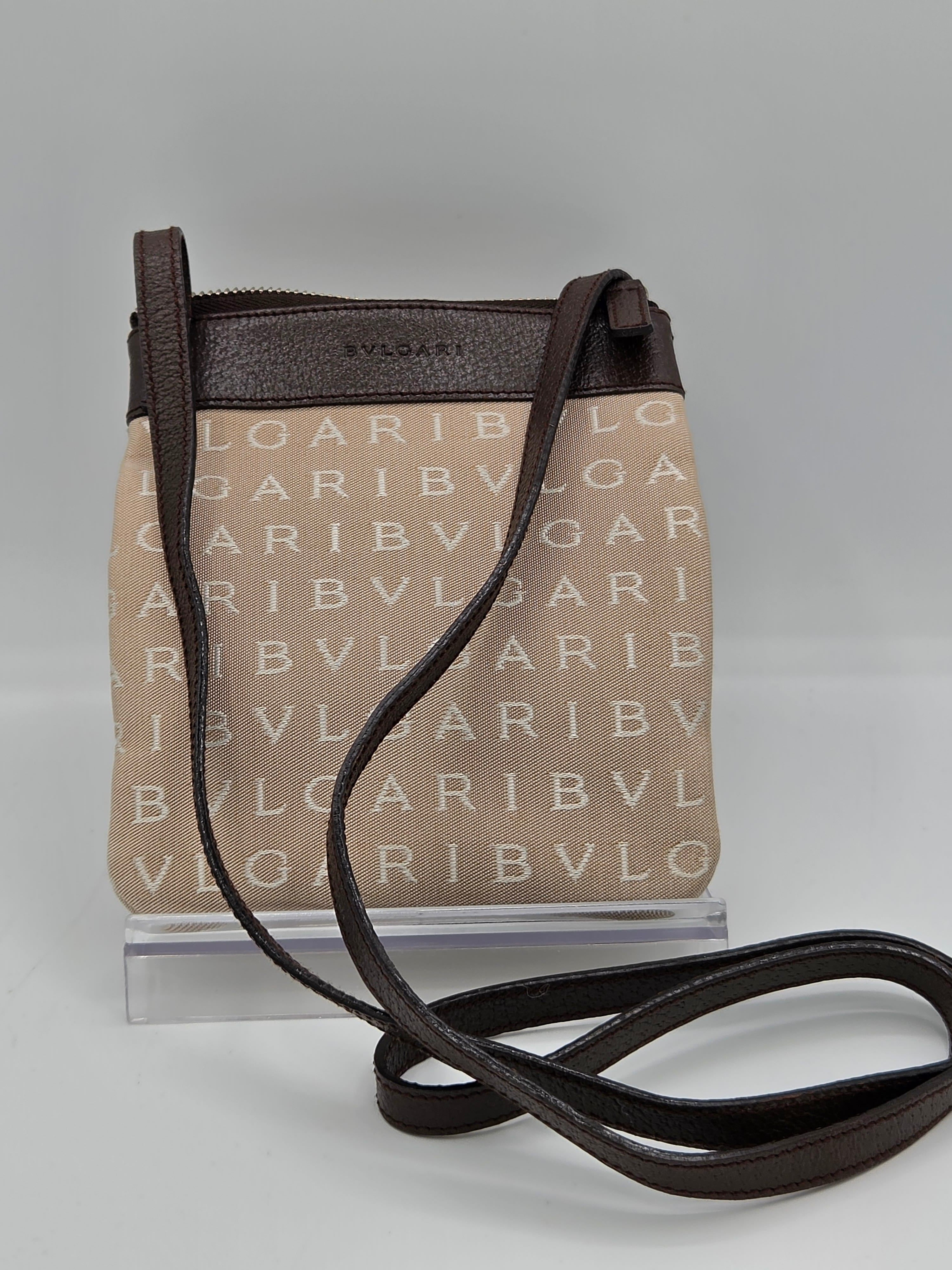 A small crossbody bag with a beige canvas body featuring a repeating 'BVLGARI' monogram pattern, dark brown leather trim and strap, and a clear plastic display insert on a light gray background.