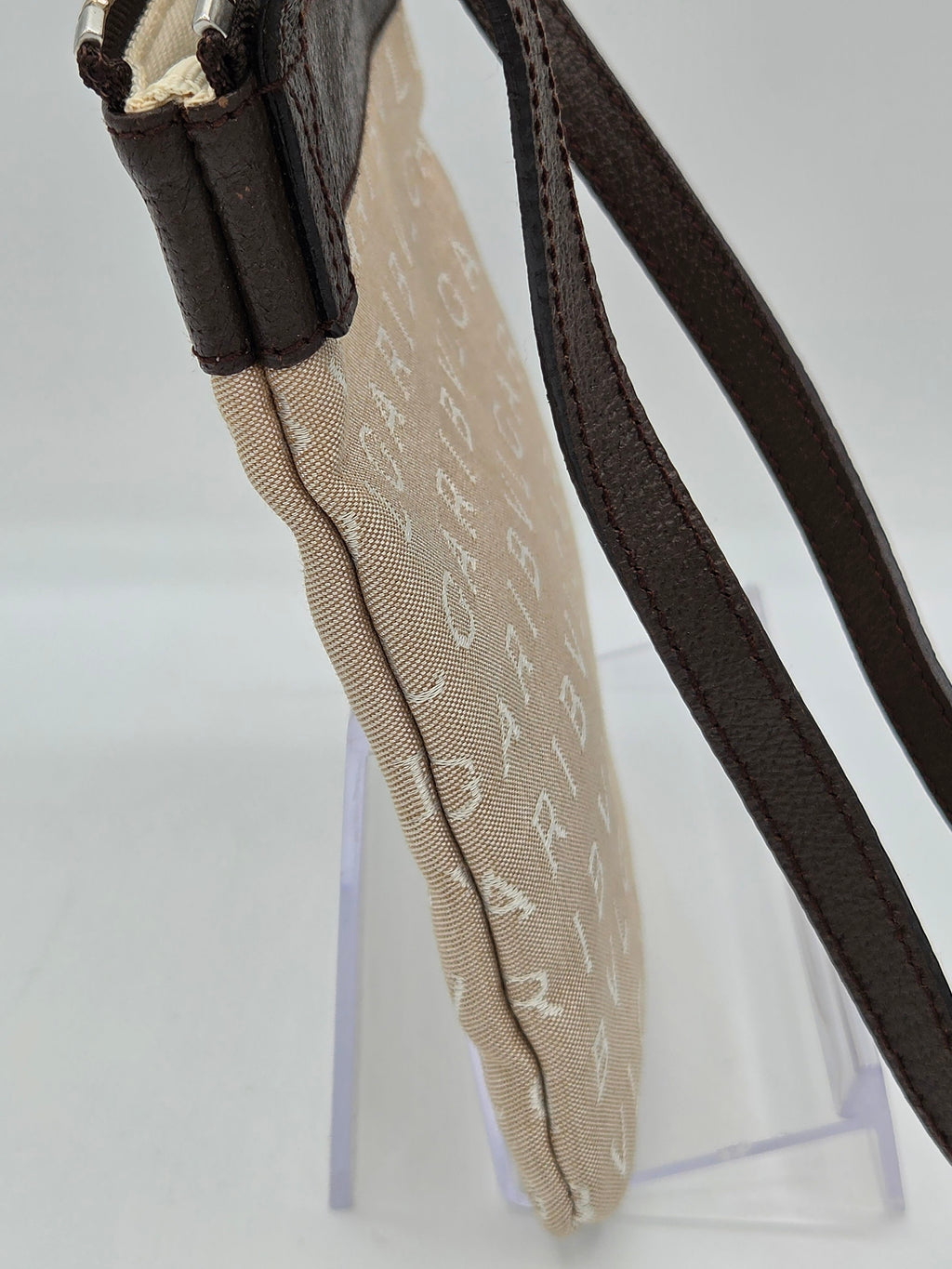 Close-up of a beige monogram canvas pouch with a dark brown leather strap, resting on a clear acrylic display stand against a white background.