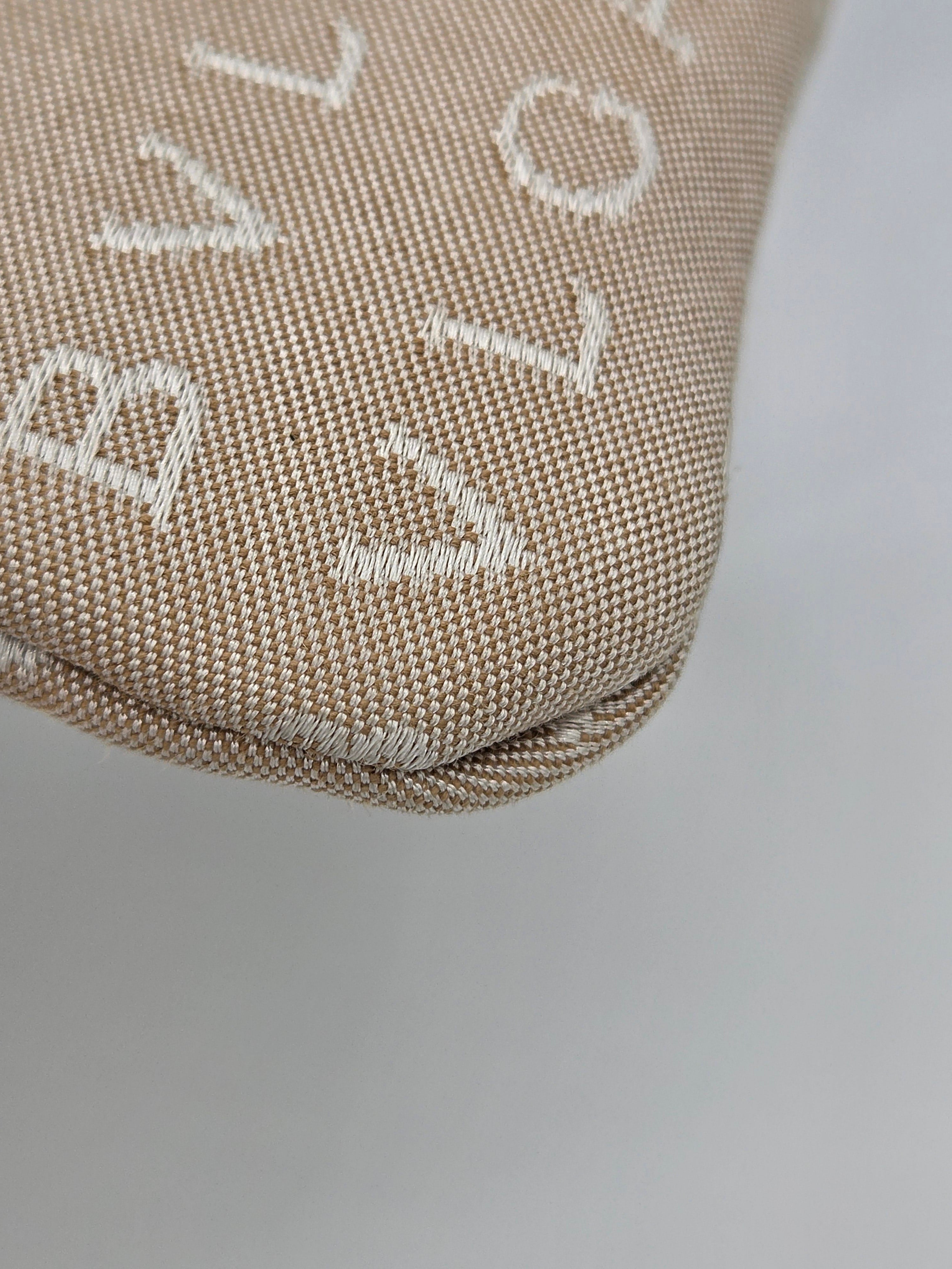 Close-up of a beige woven fabric item with white embroidered lettering, showing the texture and corner detail against a light gray background.