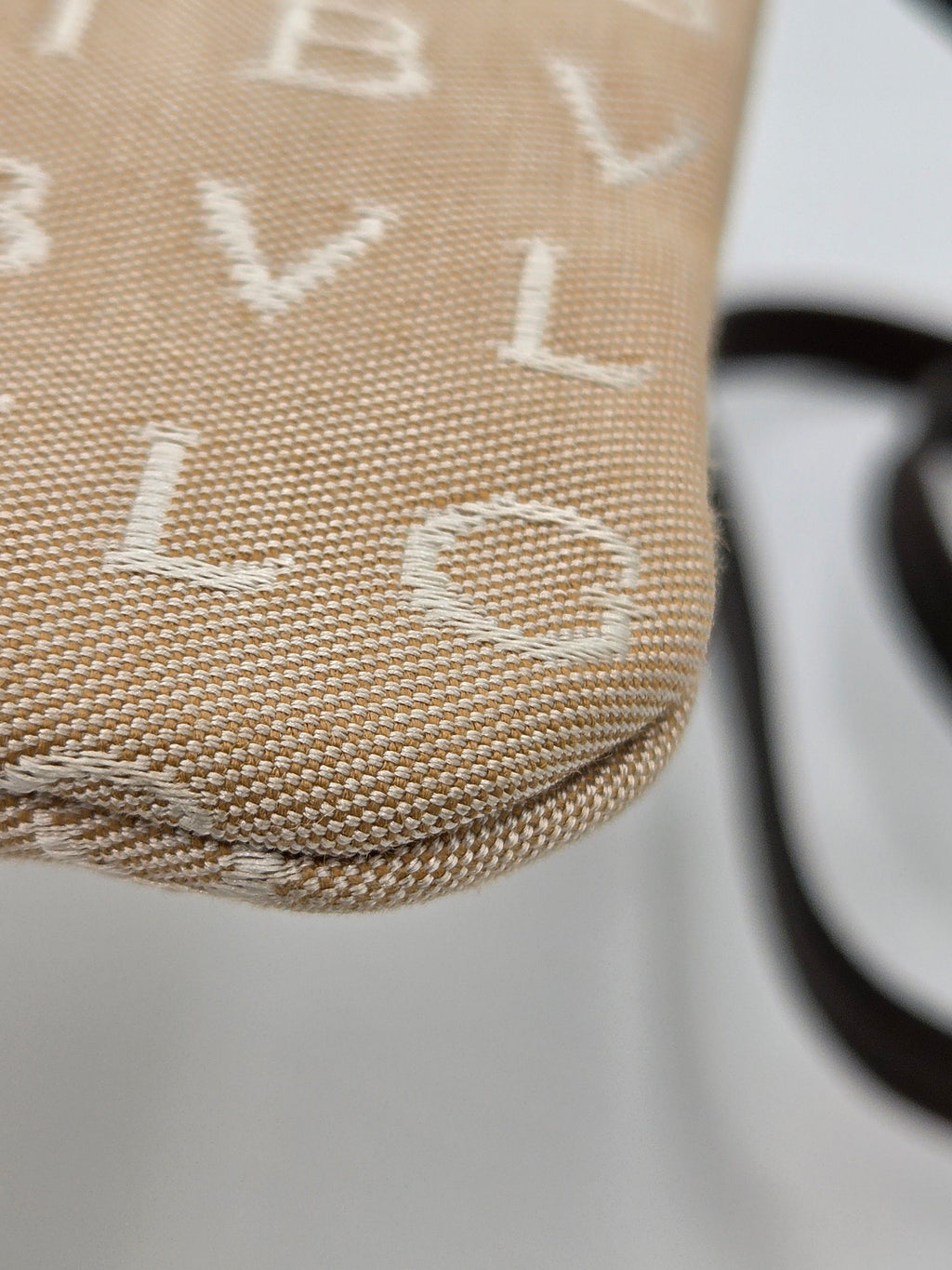 Close-up of a woven beige fabric product with white monogram letters and a dark strap on a light background.