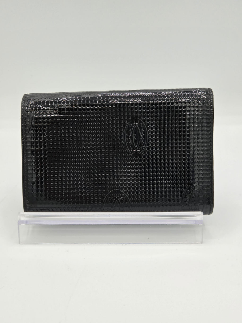 A black textured leather wallet with a crocodile pattern, featuring an embossed logo on the front, shown next to a clear plastic sleeve on a white background.
