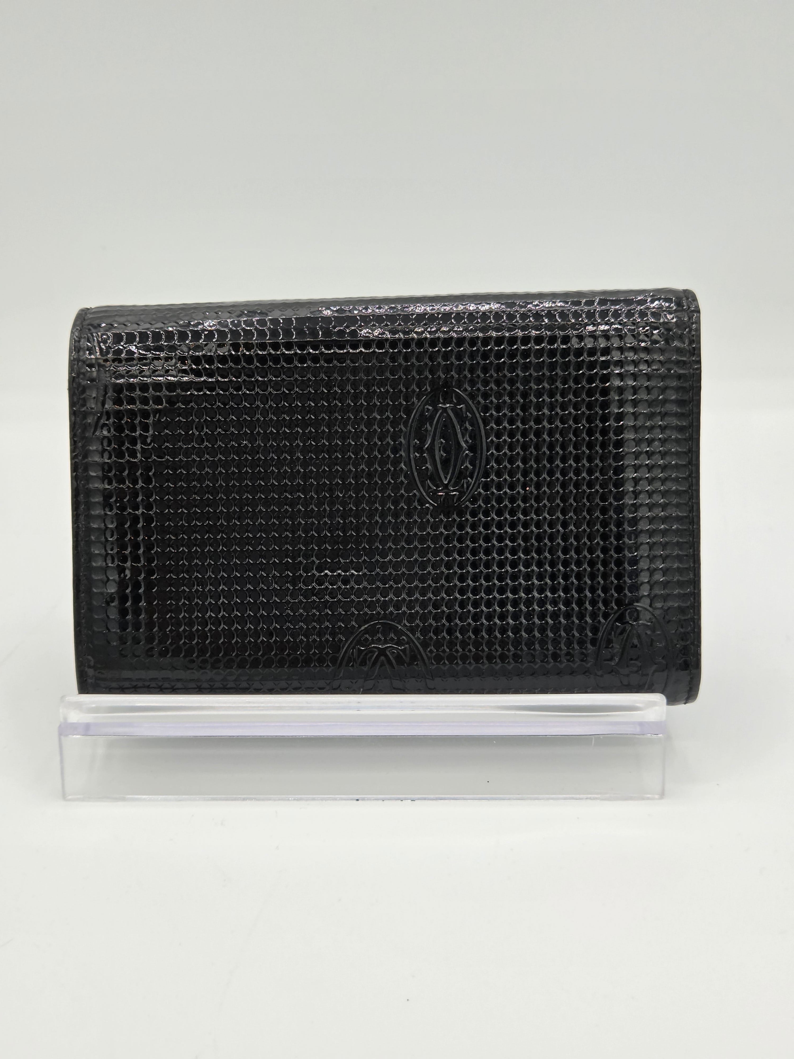 A black textured leather wallet with a crocodile pattern, featuring an embossed logo on the front, shown next to a clear plastic sleeve on a white background.