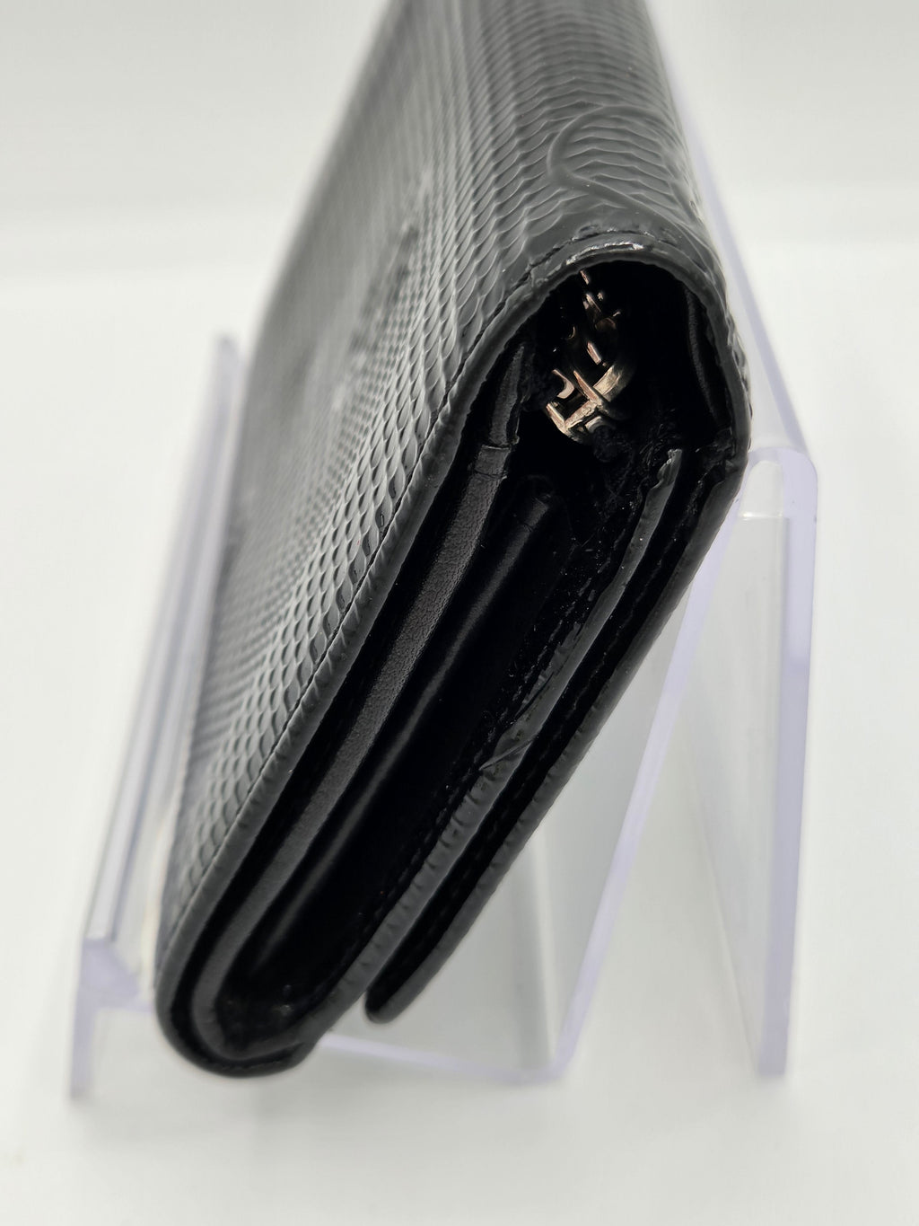 A close-up of a black textured leather wallet or notebook cover with a gold-tone metal clasp, resting on a clear acrylic display stand against a white background.