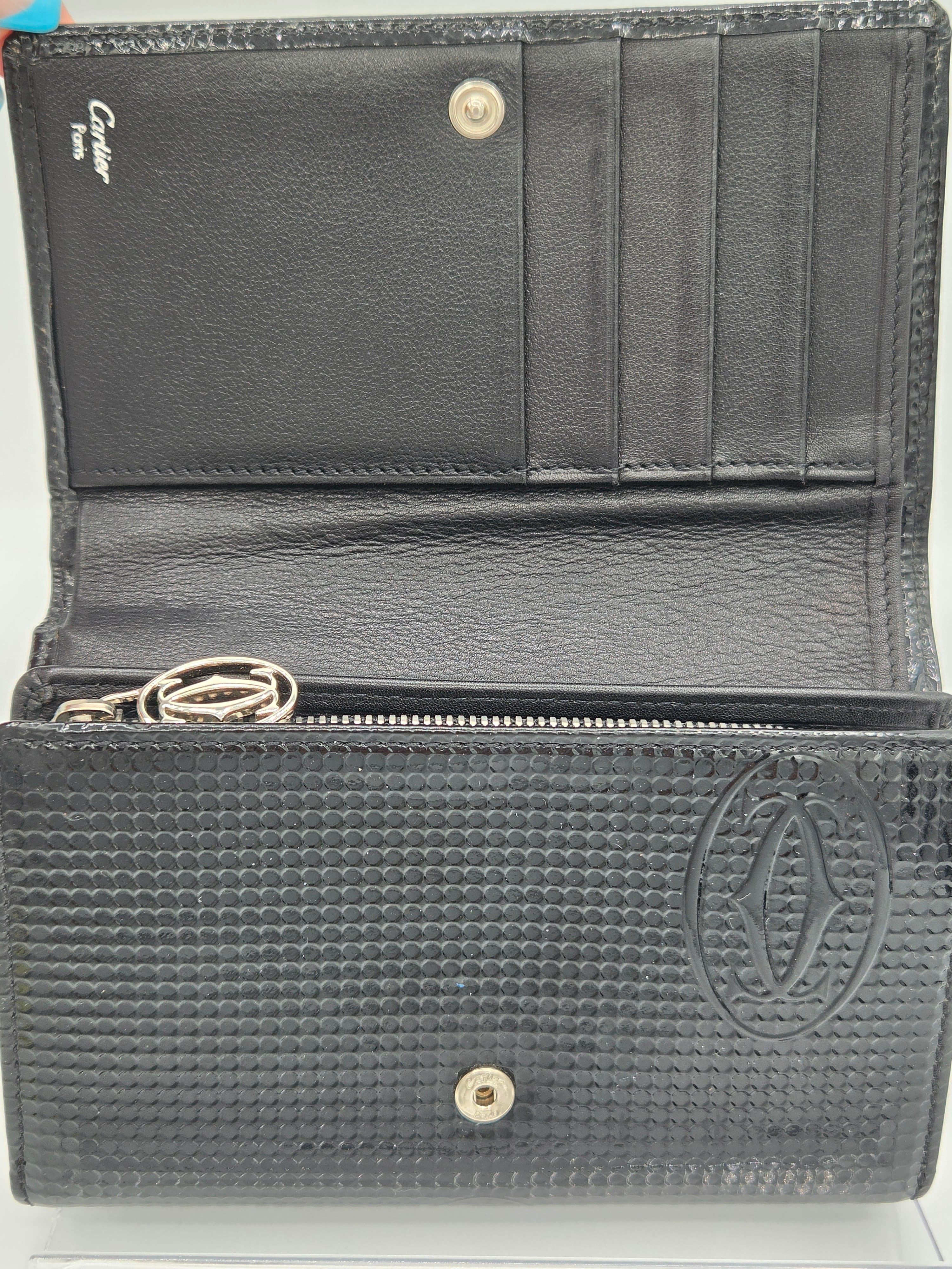A black leather wallet shown open, featuring a textured woven pattern on the exterior, card slots on the interior left, a zip pocket, and a Cartier logo printed near the bottom left.