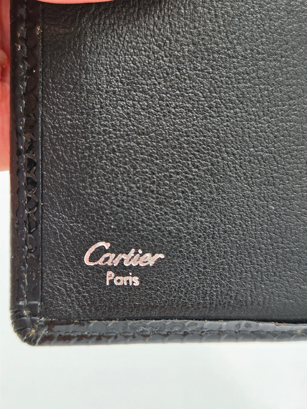 Close-up of a black textured leather wallet corner with a debossed Cartier Paris logo and visible hand holding the edge.