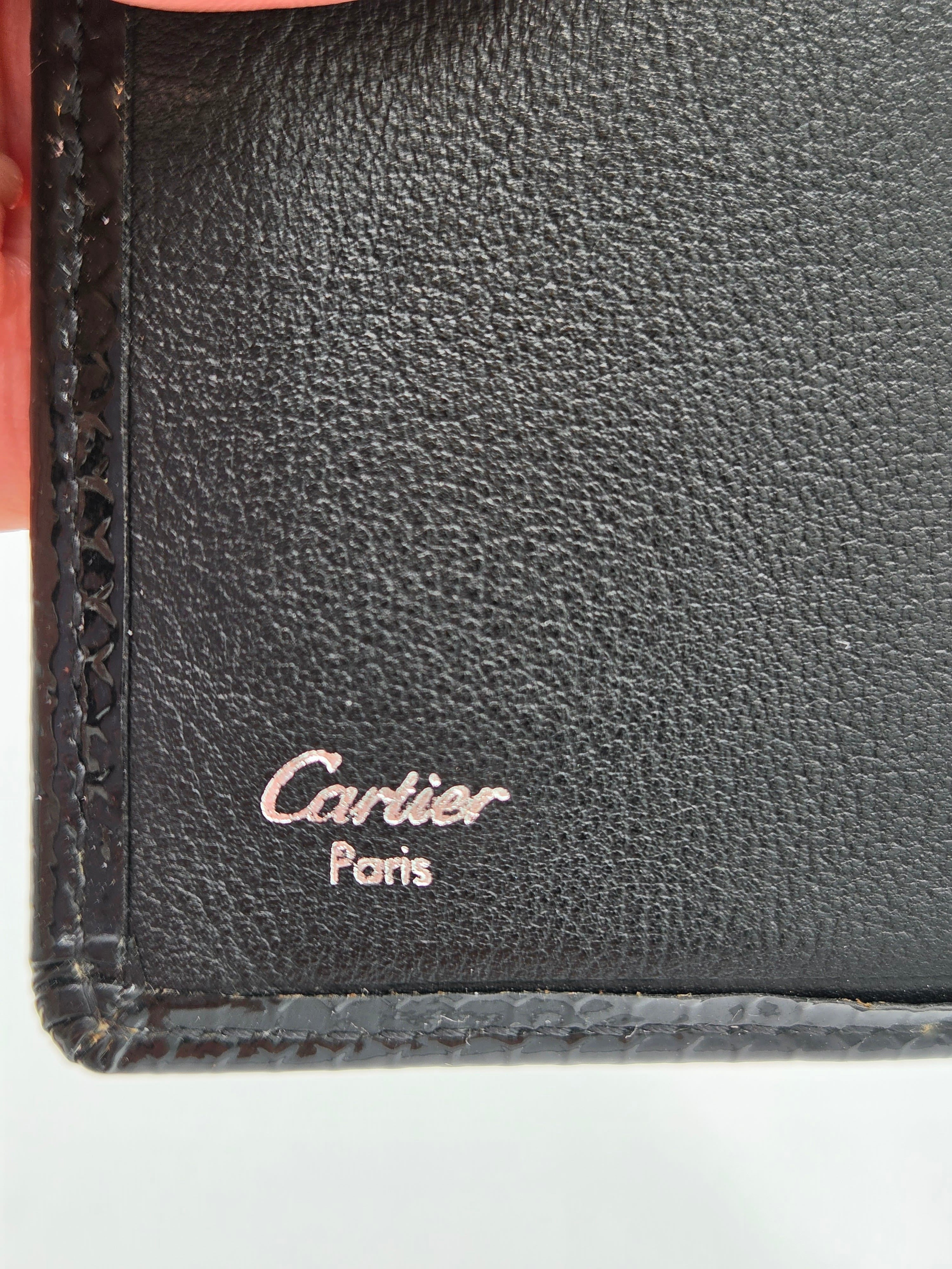 Close-up of a black textured leather wallet corner with a debossed Cartier Paris logo and visible hand holding the edge.