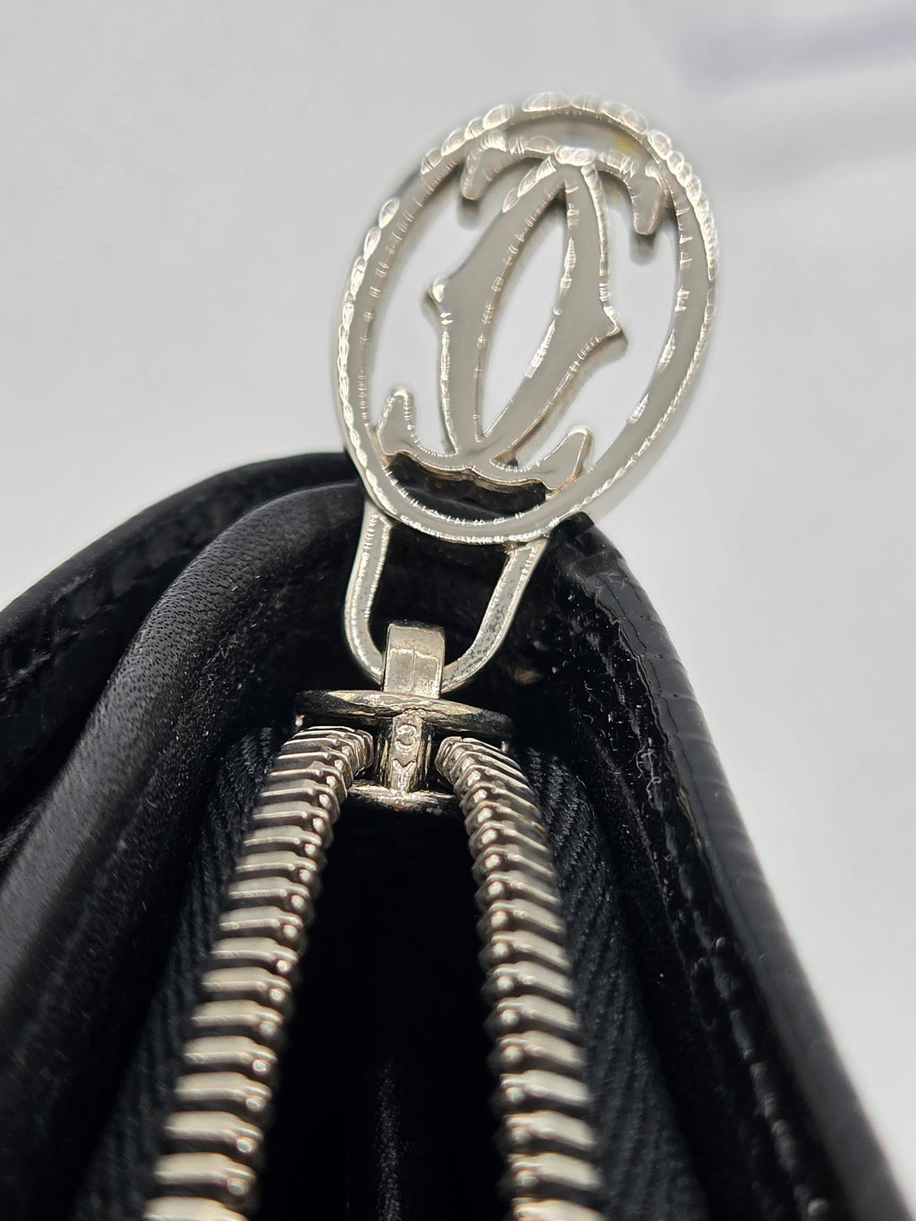 Close-up of a black leather zippered wallet or clutch featuring a silver-tone metal zipper pull with a stylized cutout logo.