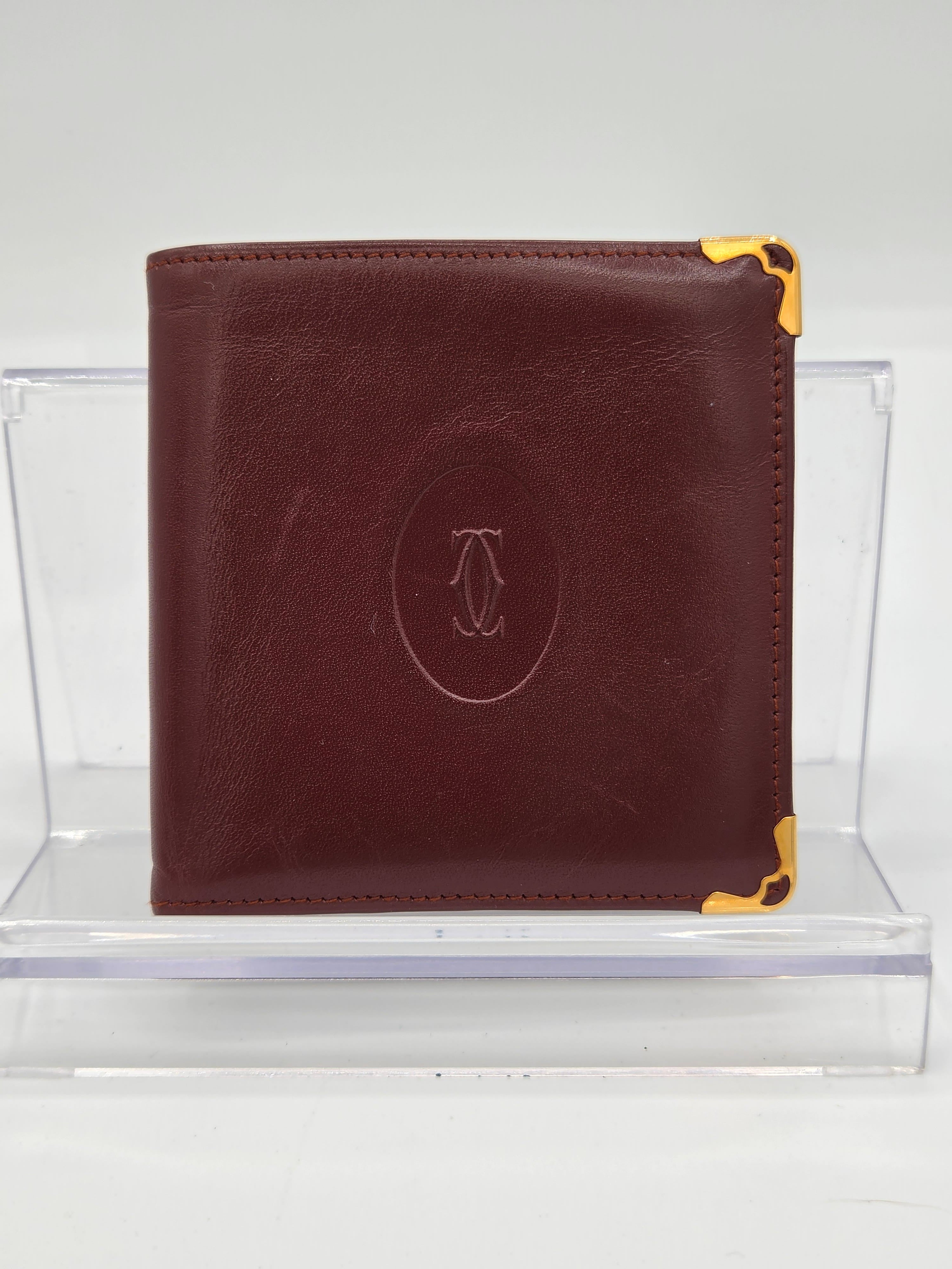 A dark brown leather bifold wallet with gold-tone metal corner protectors and an embossed oval logo on the front, displayed on a clear acrylic stand against a white background.