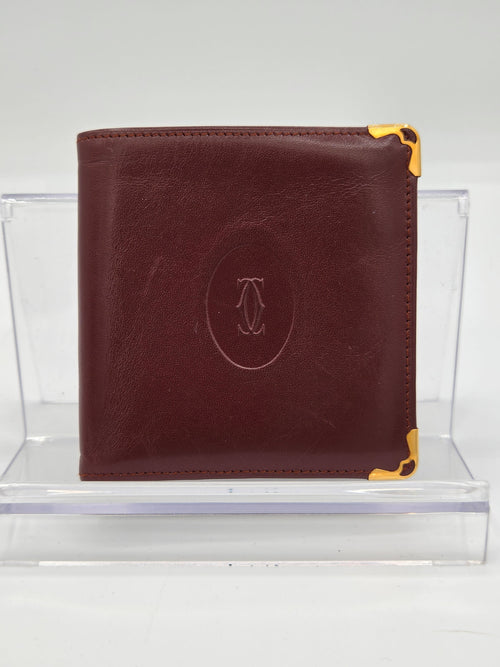 A dark brown leather bifold wallet with gold-tone metal corner protectors and an embossed oval logo on the front, displayed on a clear acrylic stand against a white background.