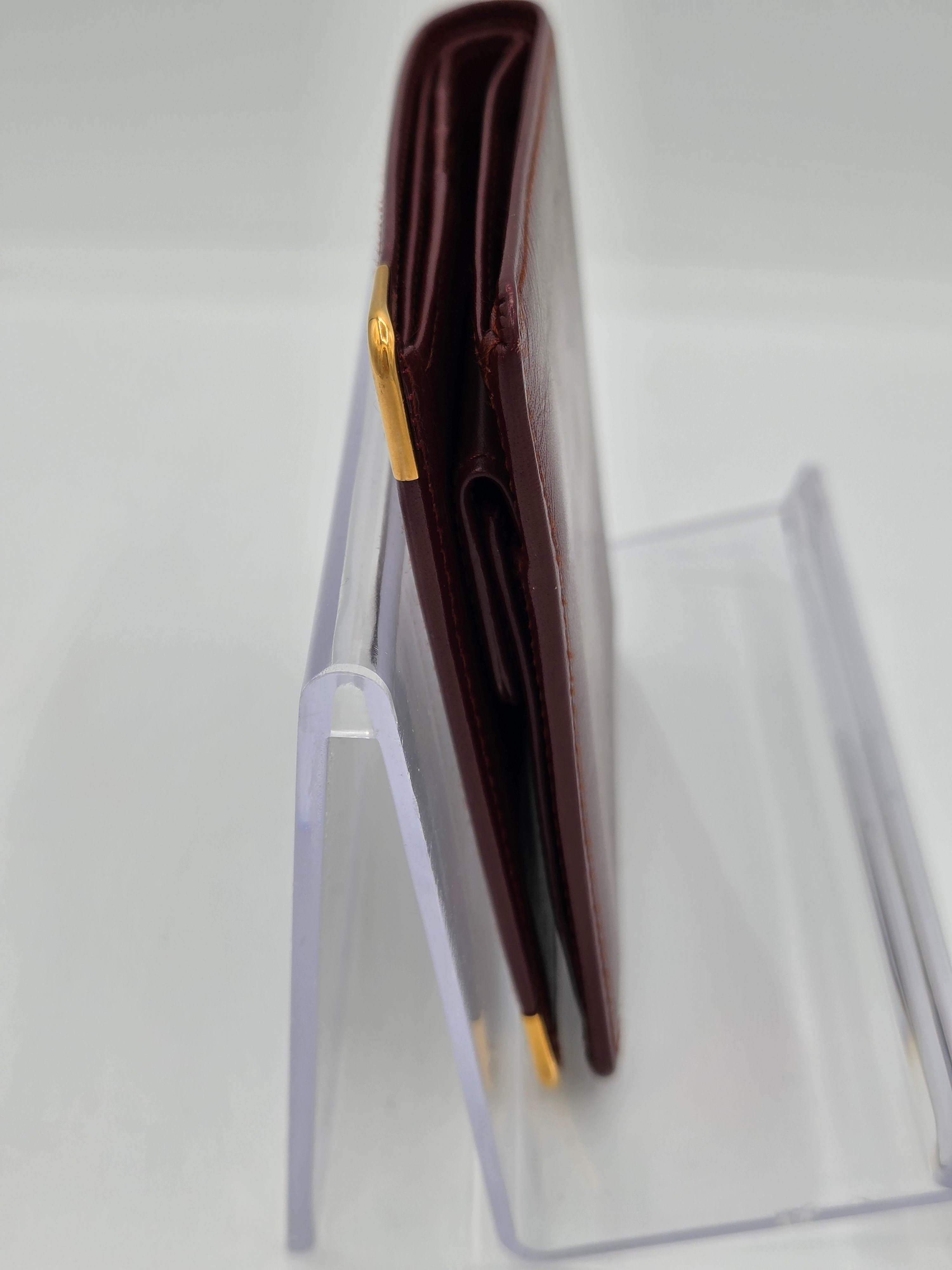 A dark brown leather wallet with gold-tone metal accents, displayed horizontally on a clear acrylic stand against a white background.