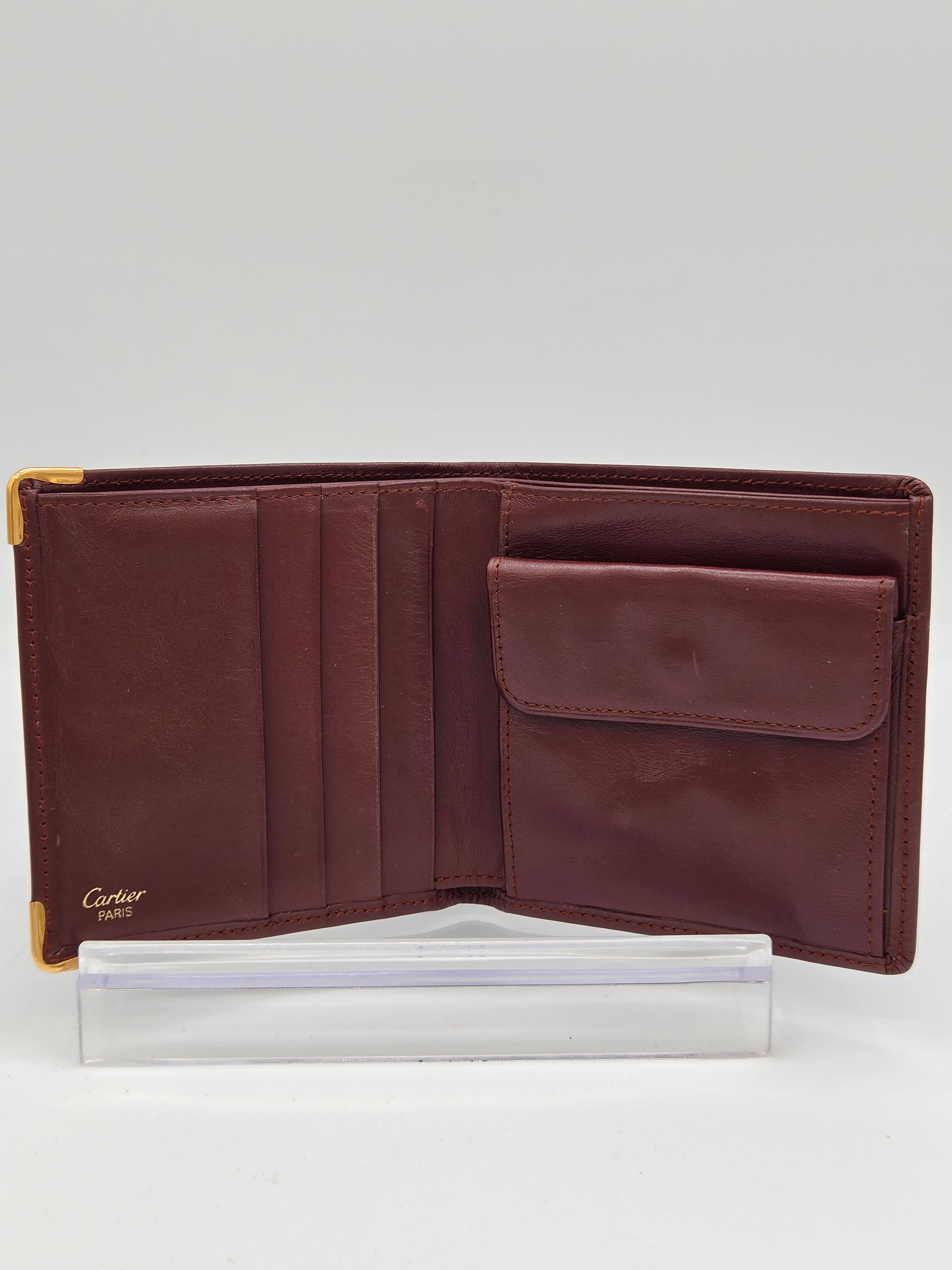 A dark brown leather bi-fold wallet shown open, displaying its interior card slots and a small gold Cartier logo in the bottom right corner, positioned next to its white paper packaging on a light gray background.