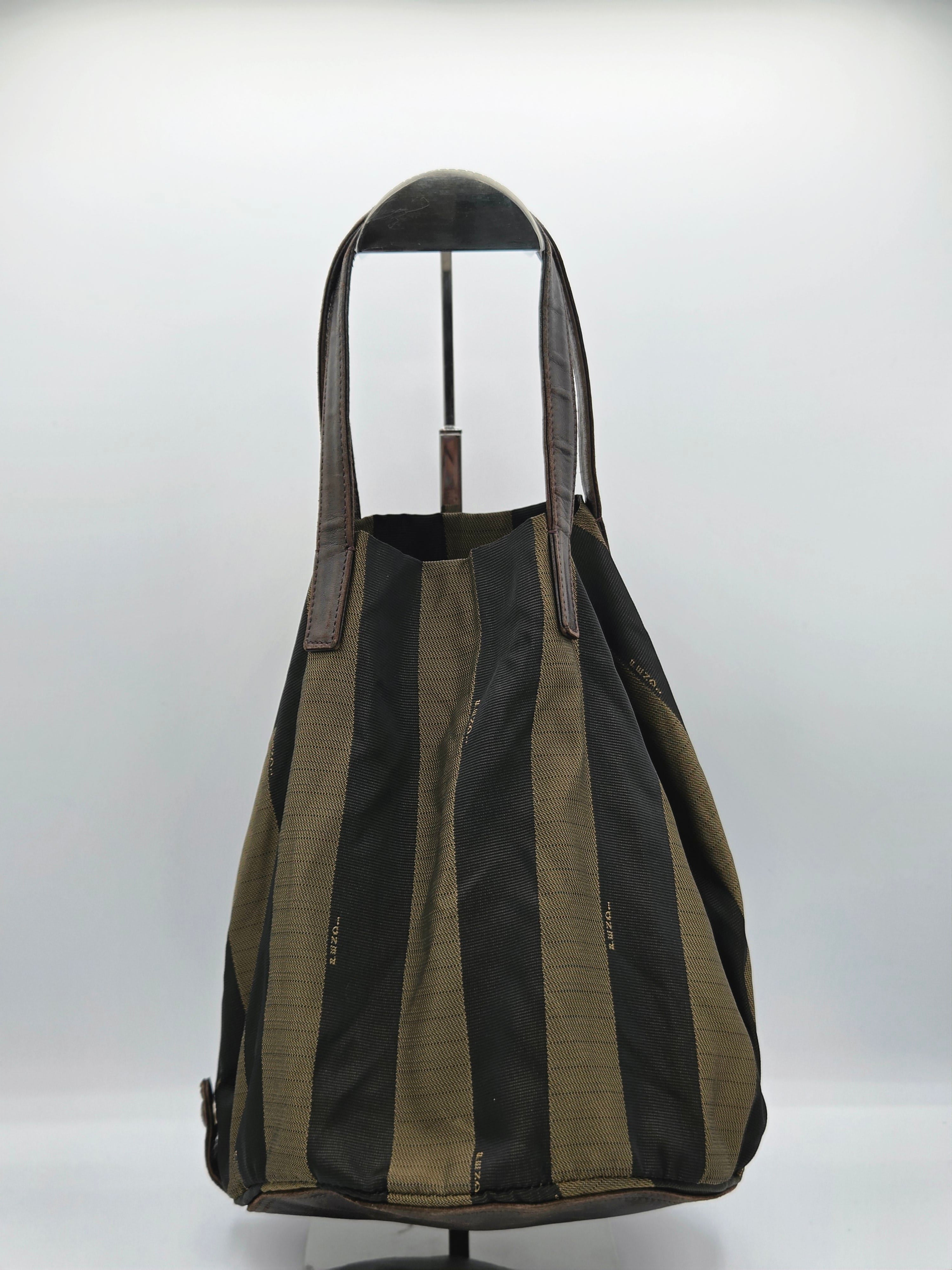 A large slouchy tote bag with vertical black and olive green stripes, displayed on a black mannequin stand against a light gray studio background.