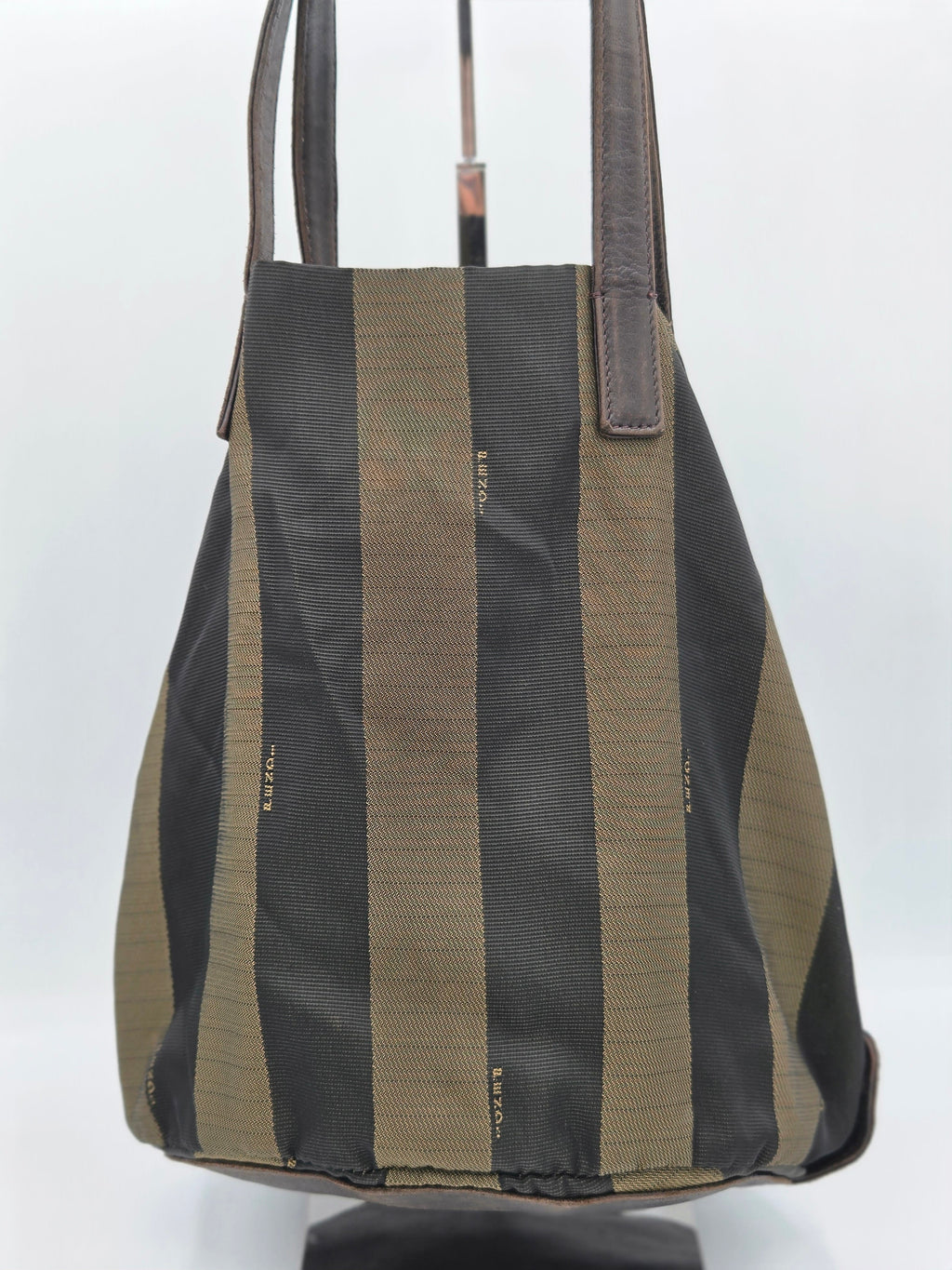 A canvas tote bag with vertical stripes in black and olive green, featuring dark brown leather shoulder straps, displayed on a stand against a white background.