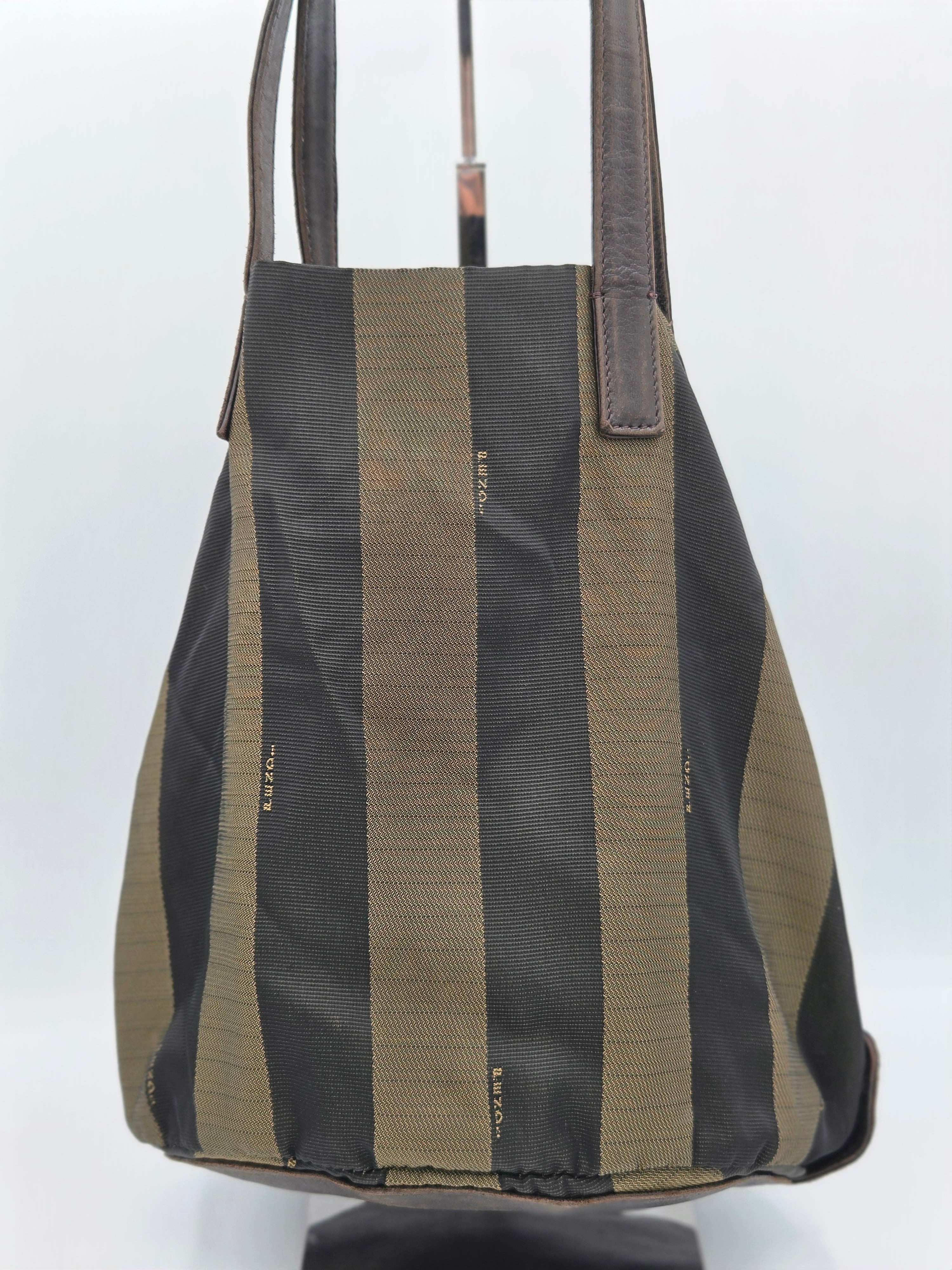 A canvas tote bag with vertical stripes in black and olive green, featuring dark brown leather shoulder straps, displayed on a stand against a white background.