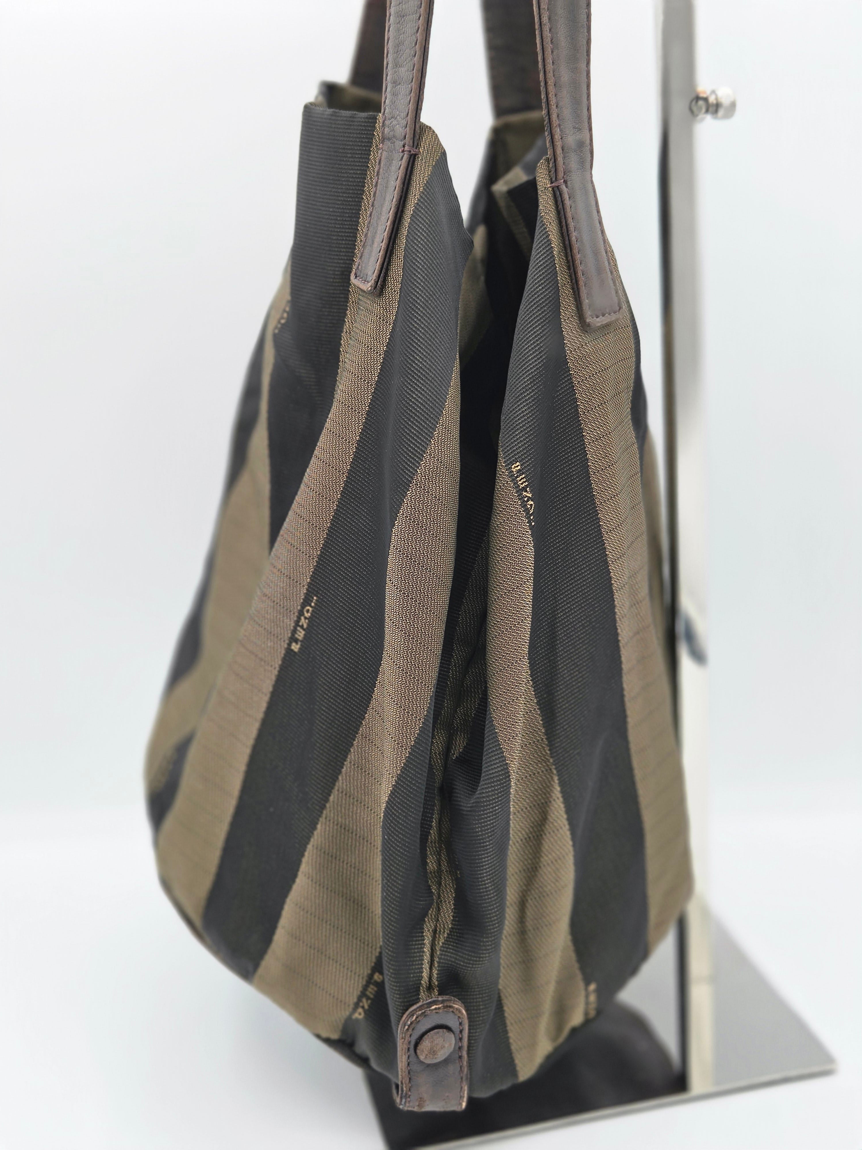 A striped canvas shoulder bag with dark gray and olive green vertical stripes, featuring brown leather handles and a small leather tab detail, displayed on a stand against a white background.