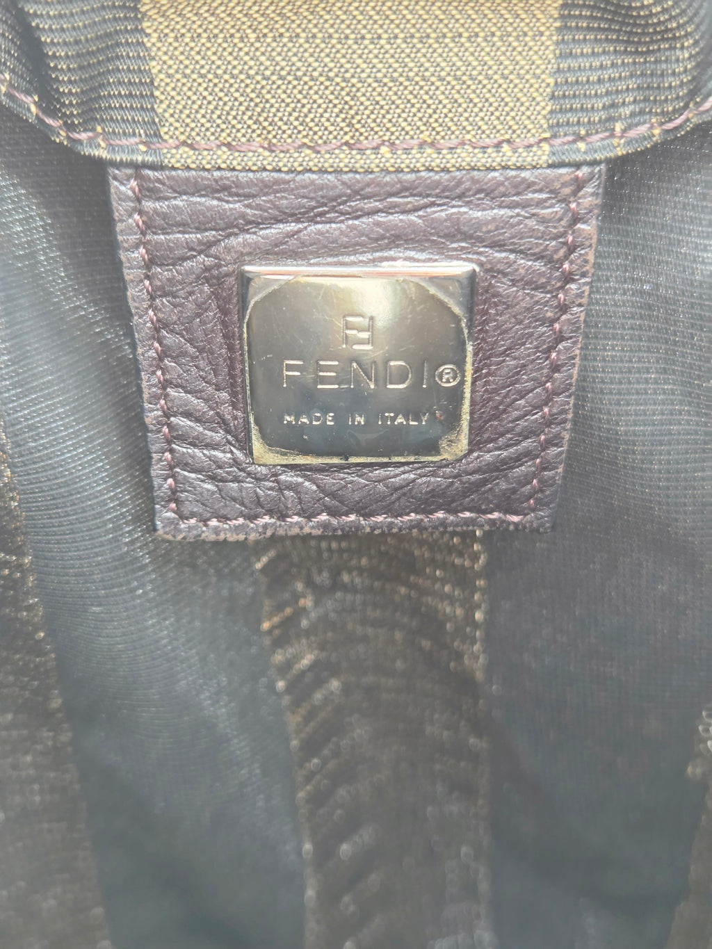 Close-up of a Fendi leather brand patch with a silver-tone metal plaque engraved with 'FENDI MADE IN ITALY' on a dark textured fabric background.
