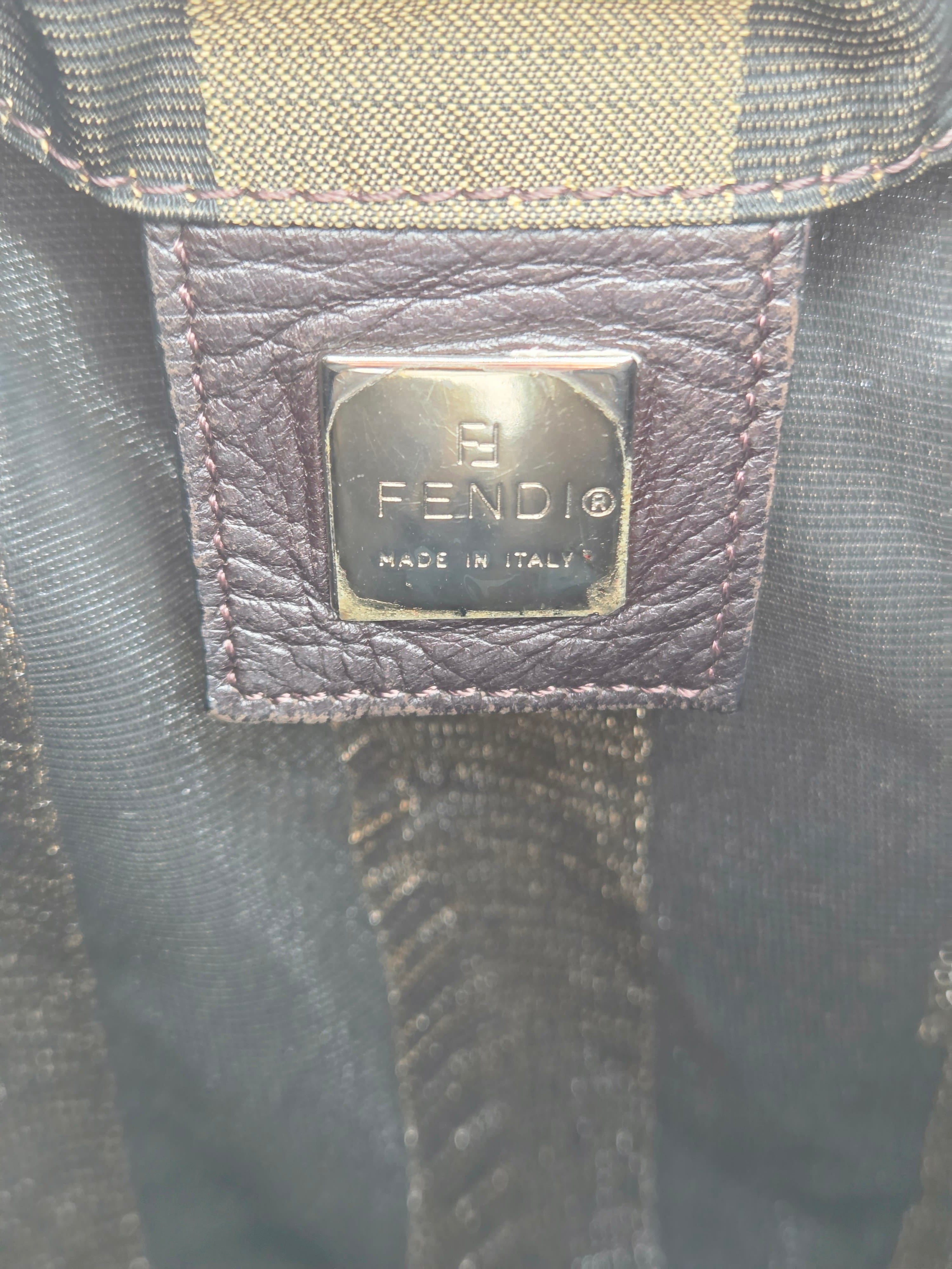 Close-up of a Fendi leather brand patch with a silver-tone metal plaque engraved with 'FENDI MADE IN ITALY' on a dark textured fabric background.