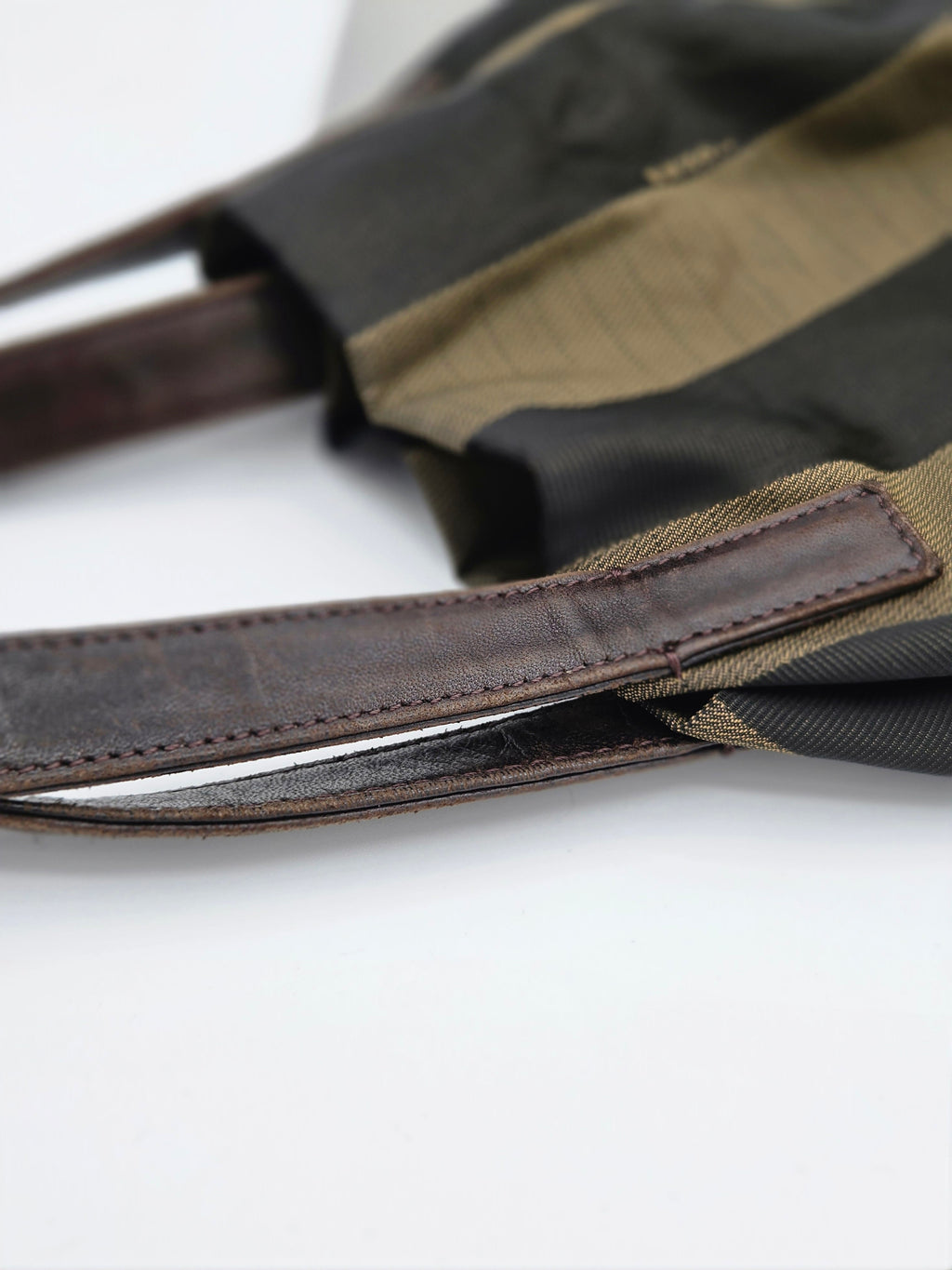 Close-up of a bag strap and fabric detail, showing dark brown leather trim with stitching over a black and olive green striped textile on a white background.
