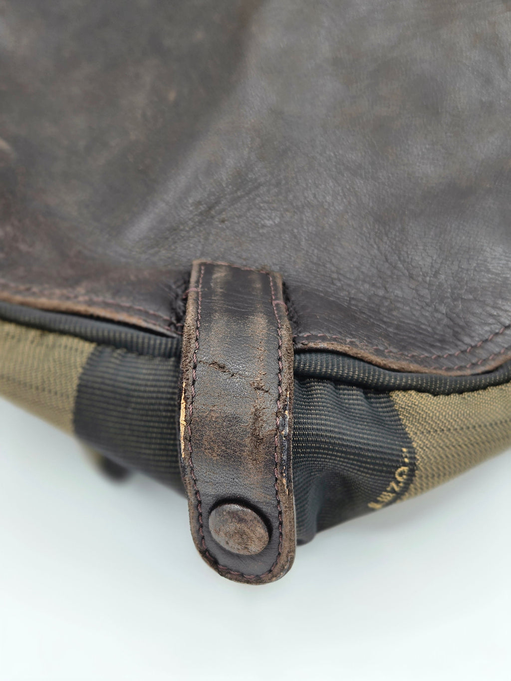 Close-up detail of a dark brown leather strap with a metal snap button closure, attached to a dark gray and olive green fabric bag against a white background.