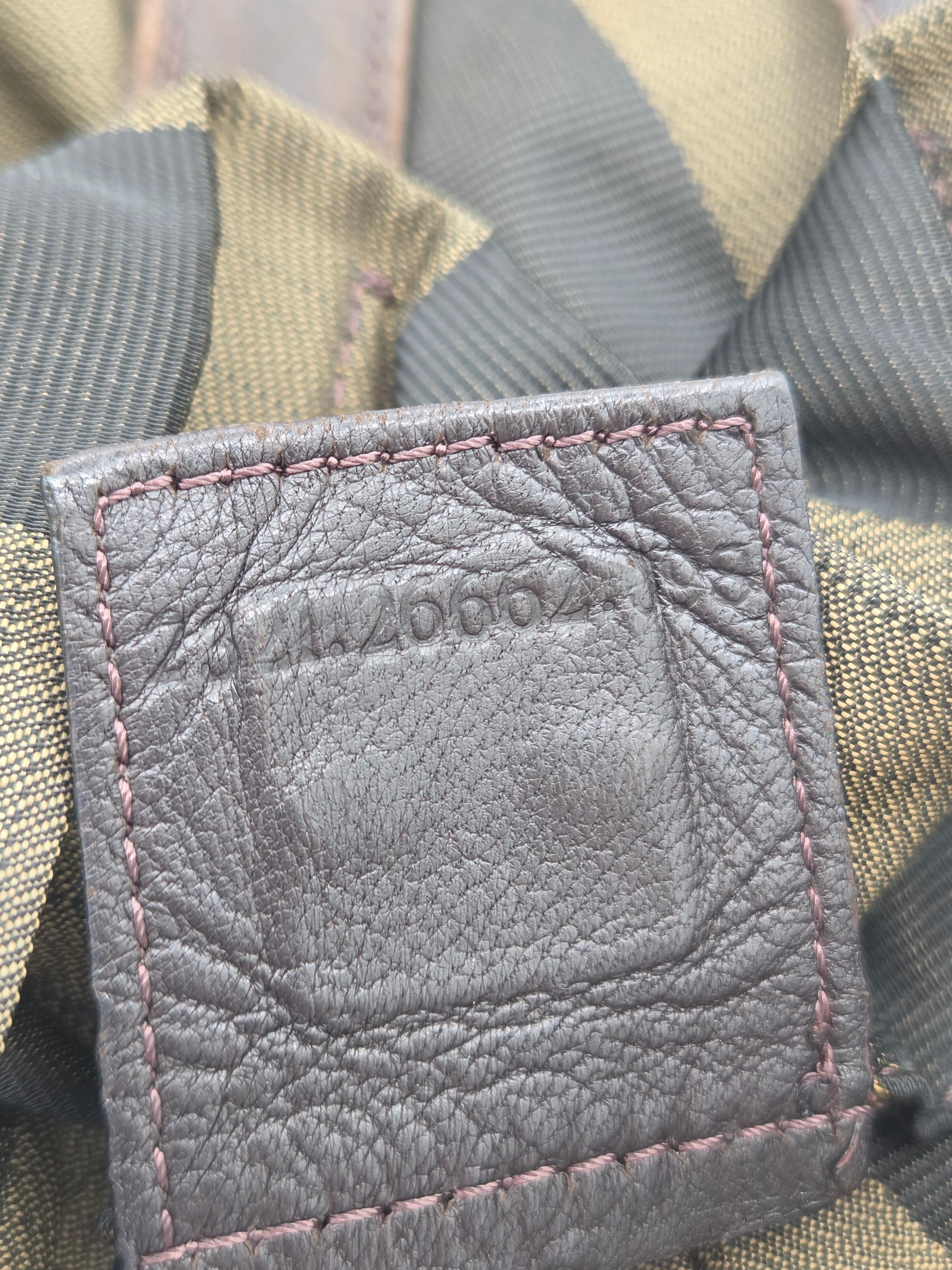 Close-up of a gray textured leather patch with pink stitching, showing an embossed logo, set against a background of olive green and gray striped fabric.