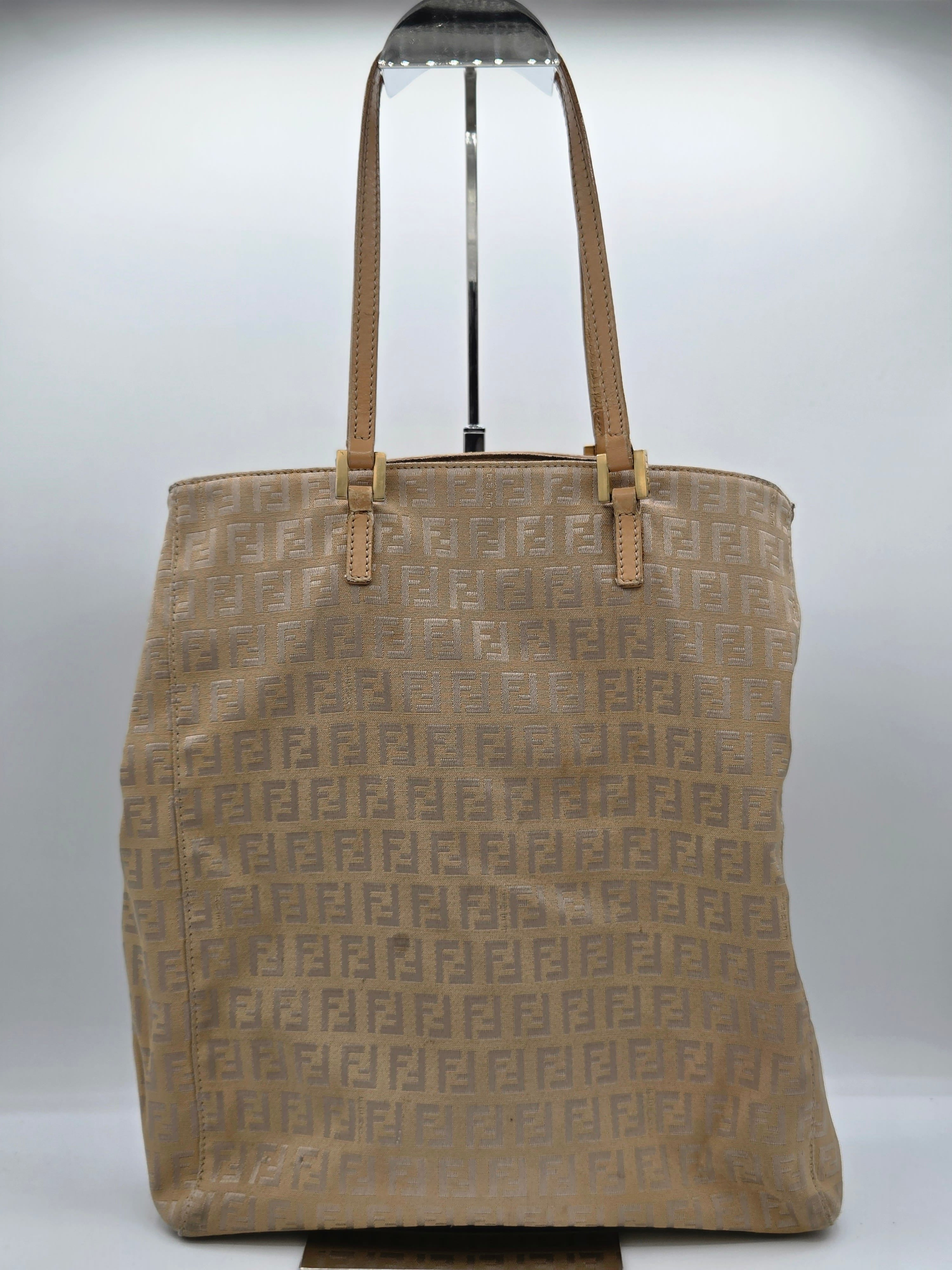 A beige Fendi tote bag with the signature Zucca monogram pattern, featuring dual shoulder straps and a black metal display stand, shown against a plain light gray background.