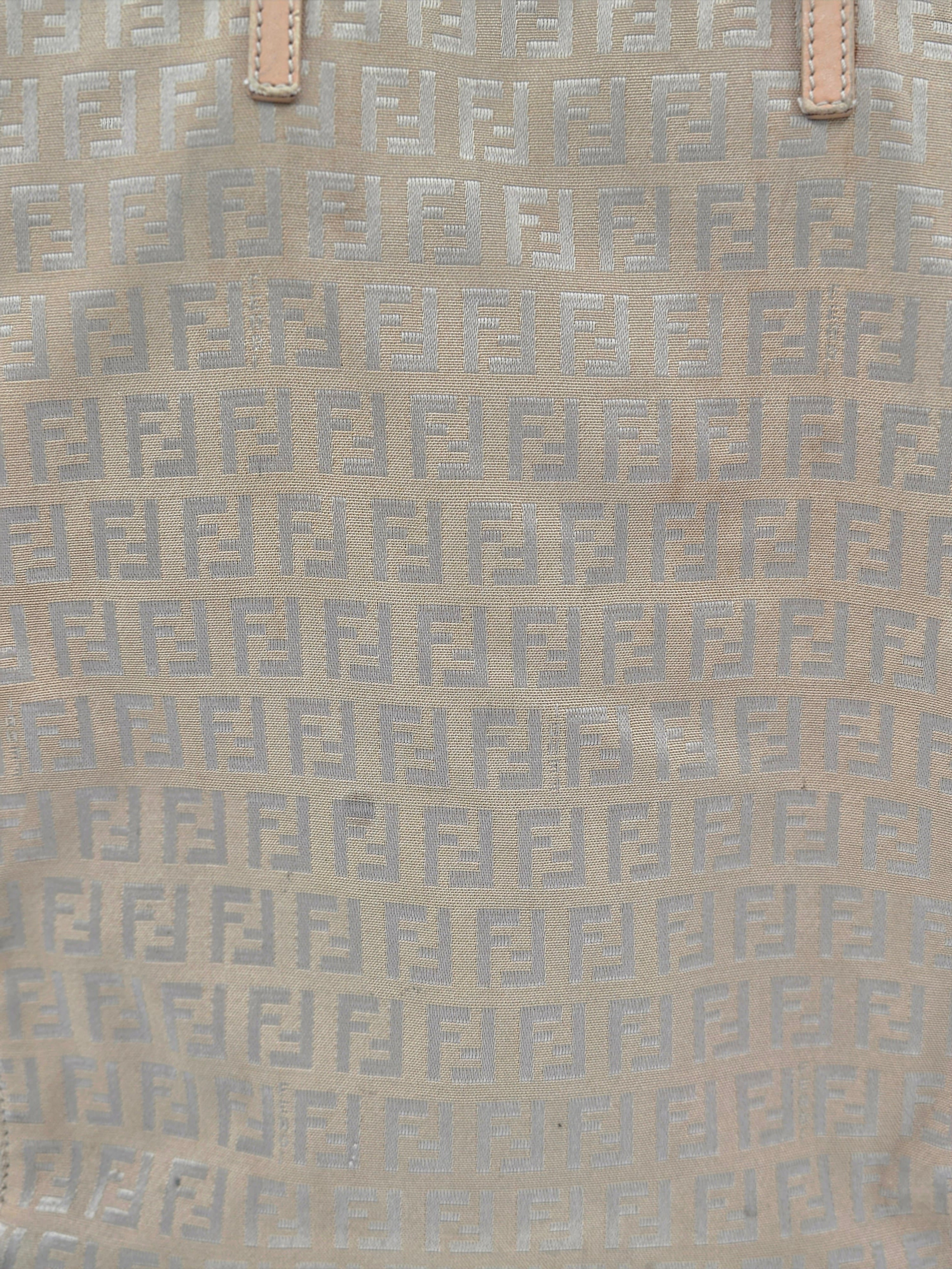 Close-up of a beige fabric with a repeating light gray geometric monogram pattern and small tan leather tab details at the top.